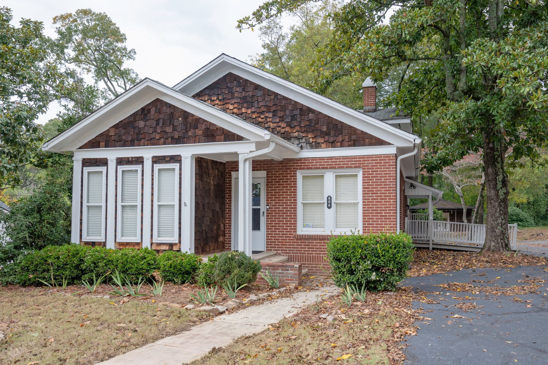 Brick house with brown shake siding, white trim, and a small porch. Green bushes and trees surround.