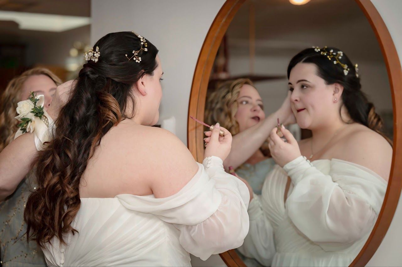 Bride looking in mirror, having makeup touched up by woman. Another woman holds a bouquet.