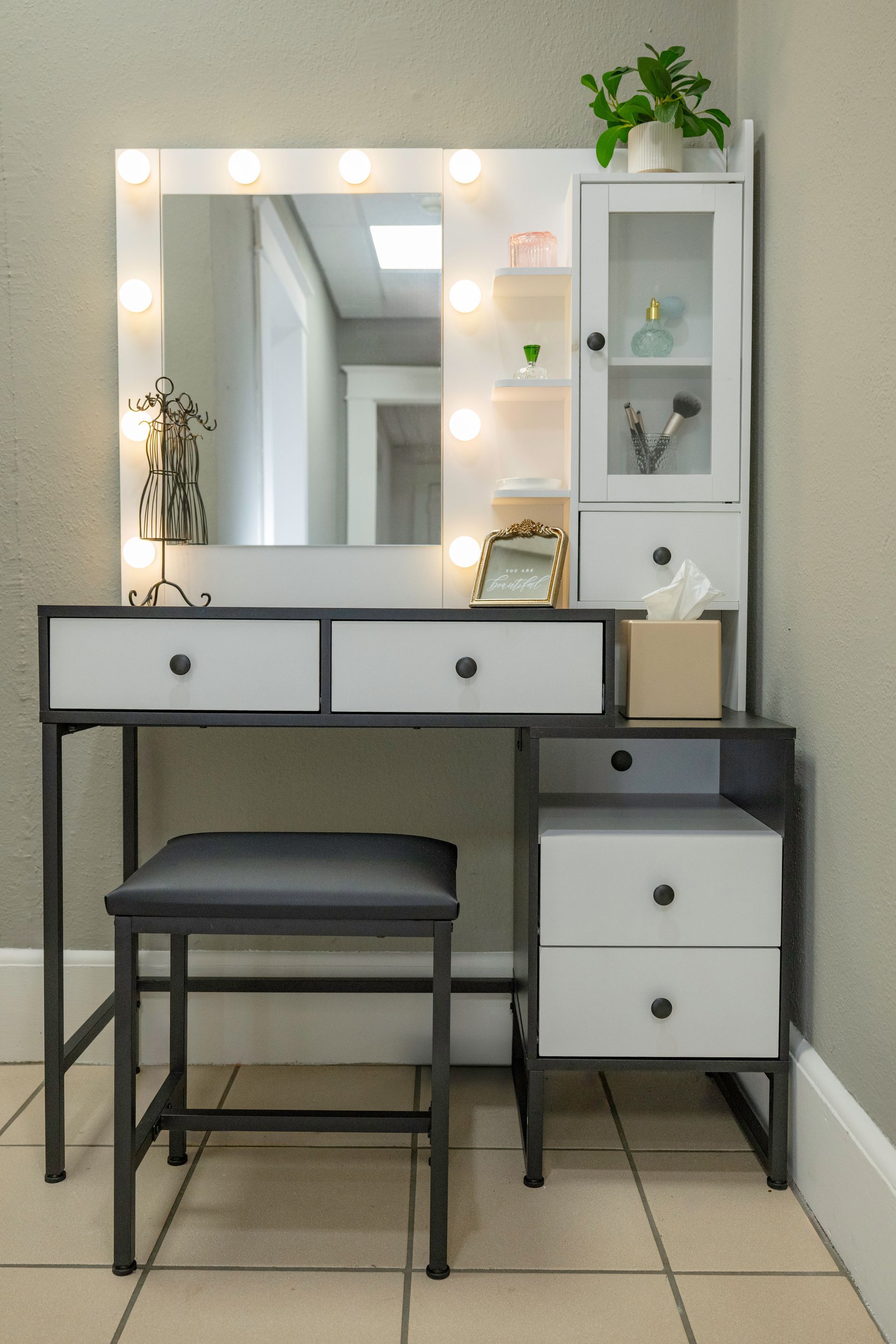 Vanity with lit mirror, drawers, stool, and storage cabinet, against a light wall.