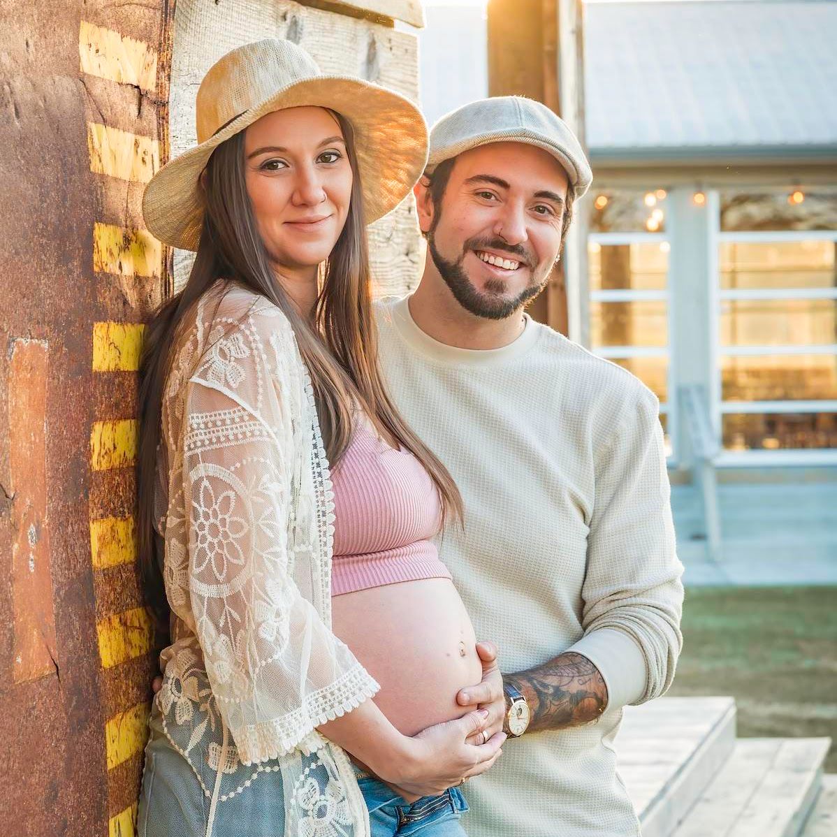 Pregnant couple posing outdoors; woman in hat, man in cap.