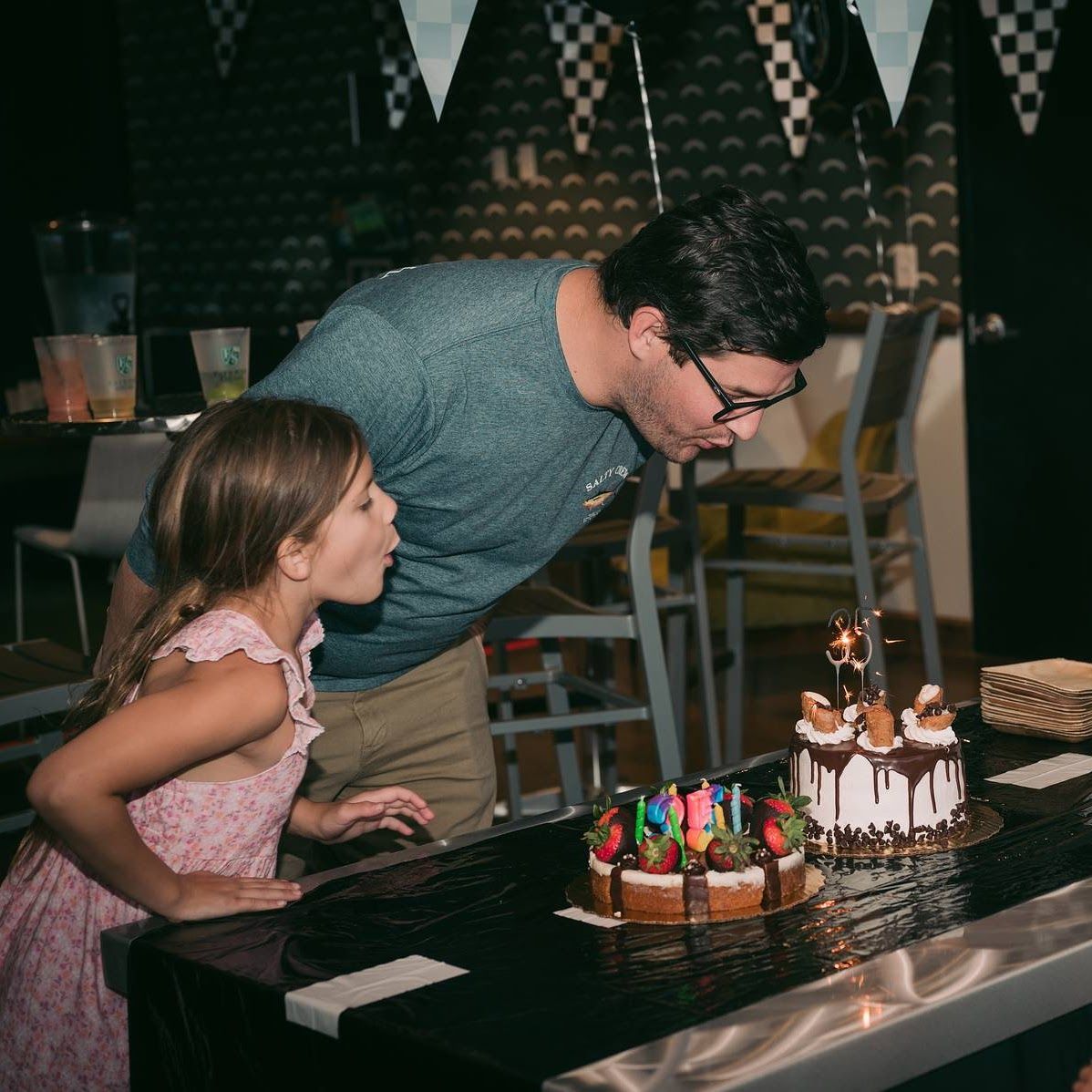 A girl and a man blowing out candles on two birthday cakes.