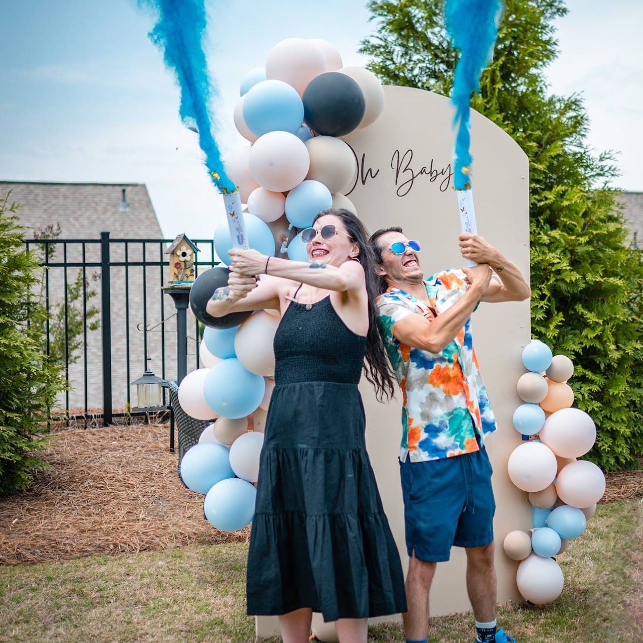 Couple holding blue smoke cannons at a gender reveal, with balloons and an 