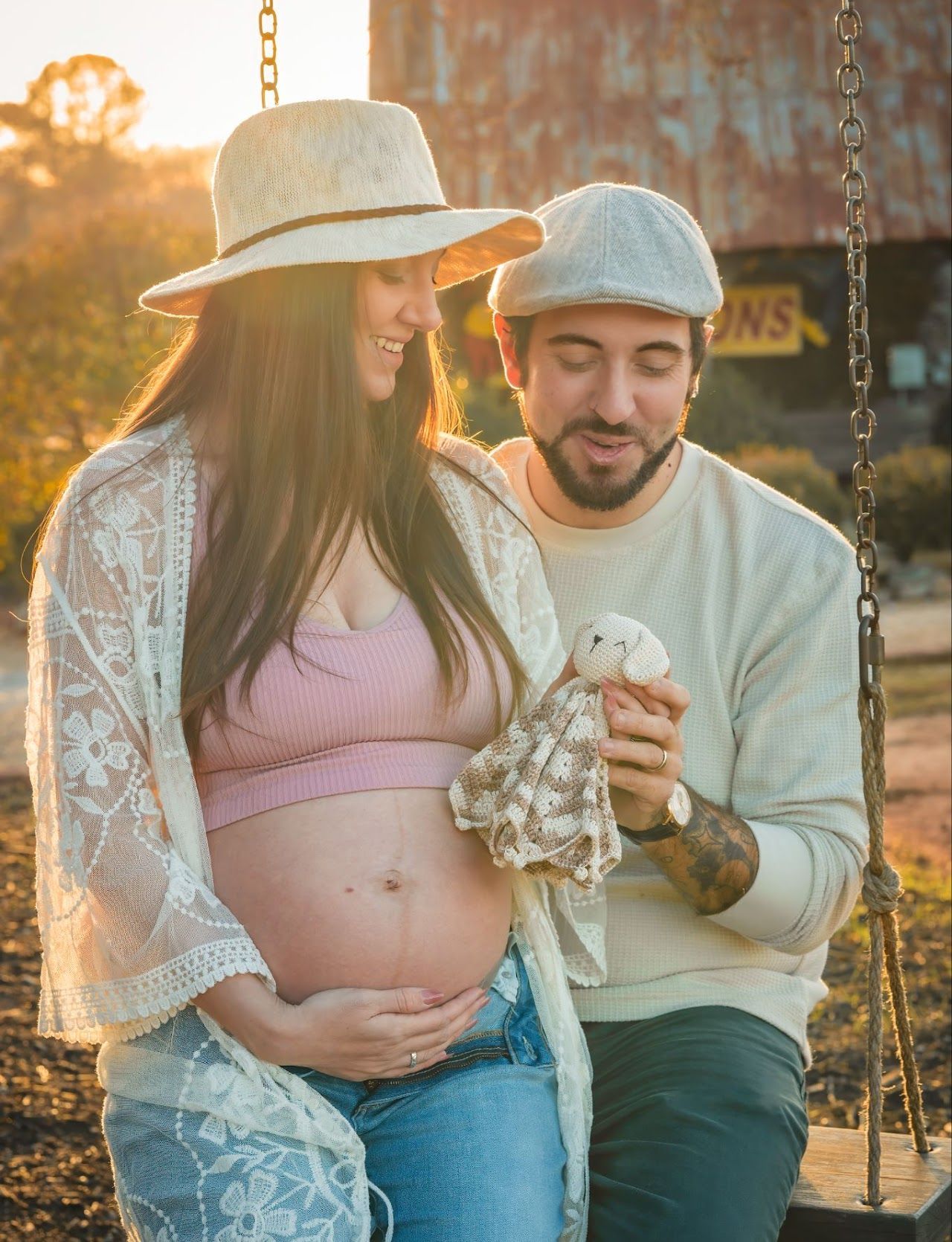 Pregnant woman and partner on a swing holding stuffed animal; outdoors, sunset, wearing hats, smiling.