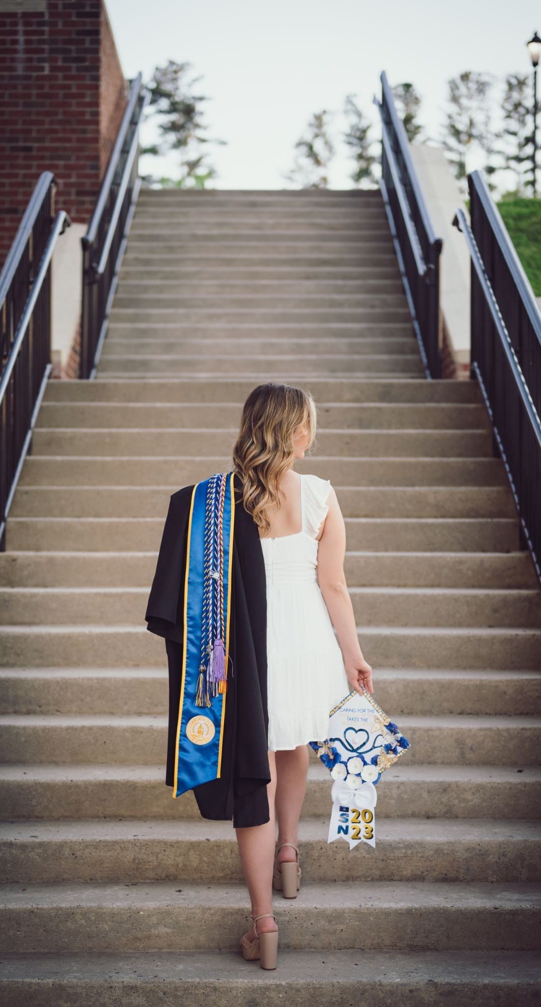Woman in white dress ascends stairs, holding graduation memorabilia and jacket.