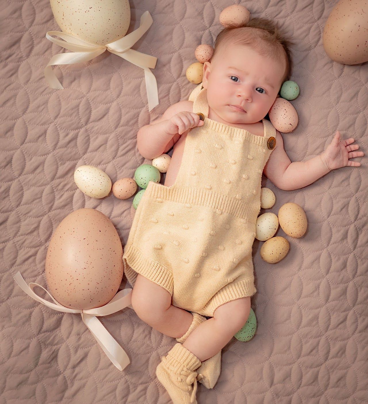 Baby in yellow romper surrounded by Easter eggs on a quilted blanket, looking at the viewer.