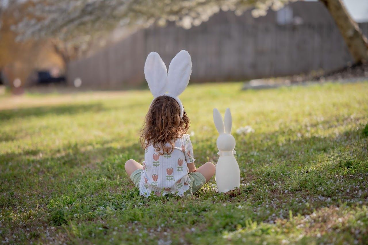 Child with bunny ears sits on grass beside a white bunny statue under a flowering tree.