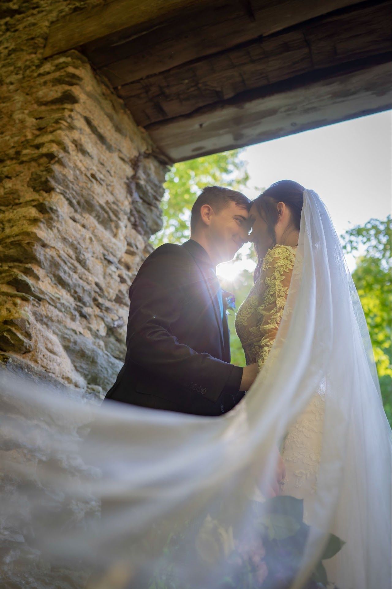 Newlyweds face each other, nearly kissing, under a stone archway. The bride's veil flows.