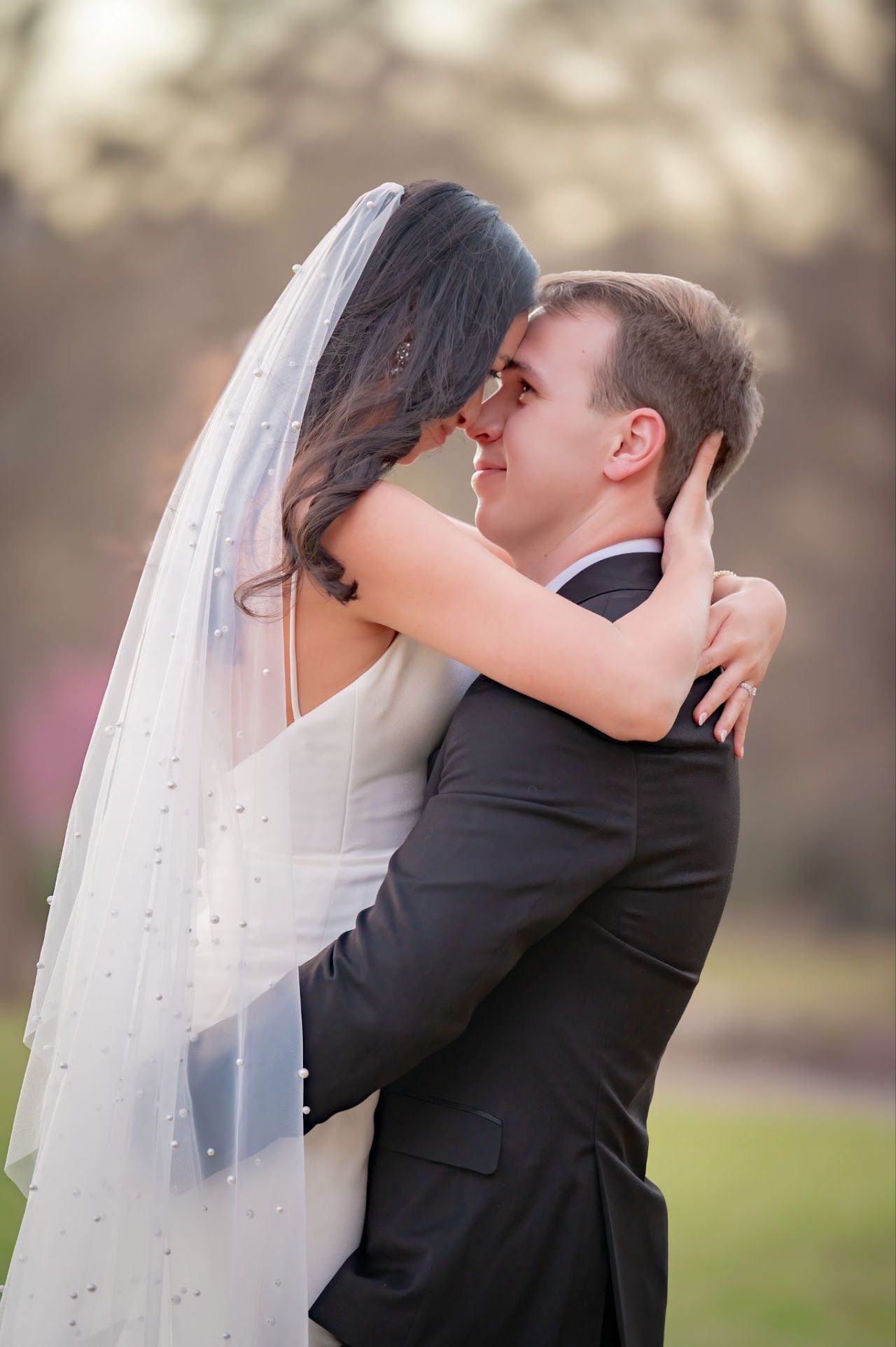 Groom holding bride, both smiling and embracing outdoors; she wears a veil and white dress, he wears a black suit.