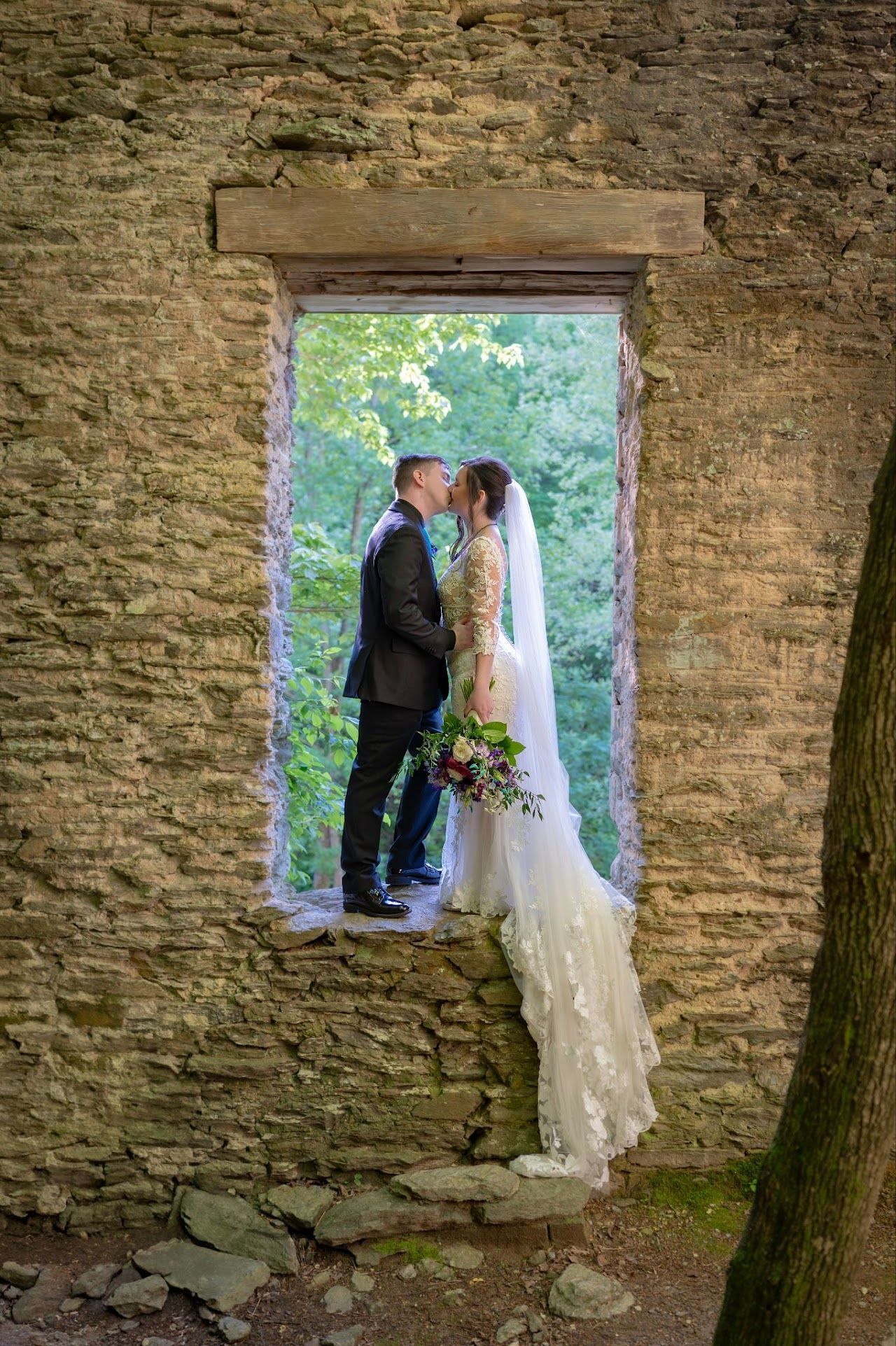 Bride and groom kissing in stone window frame, surrounded by greenery.
