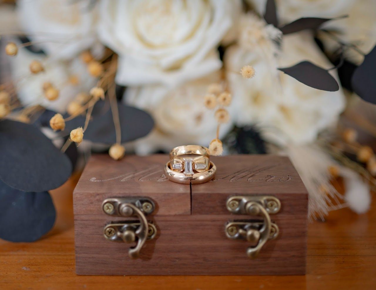 Wedding rings on a wooden box, with a blurred bouquet of white flowers in the background.