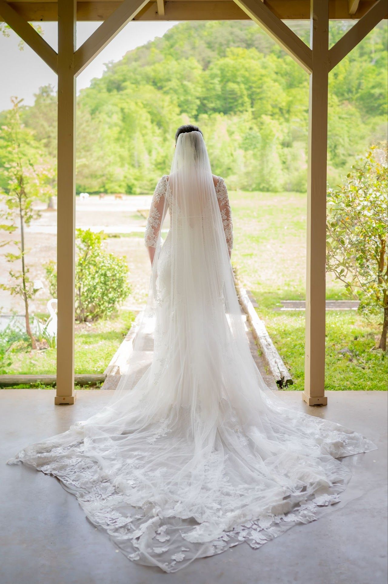 Bride in white lace gown and veil, standing on a porch, looking out at a green landscape.