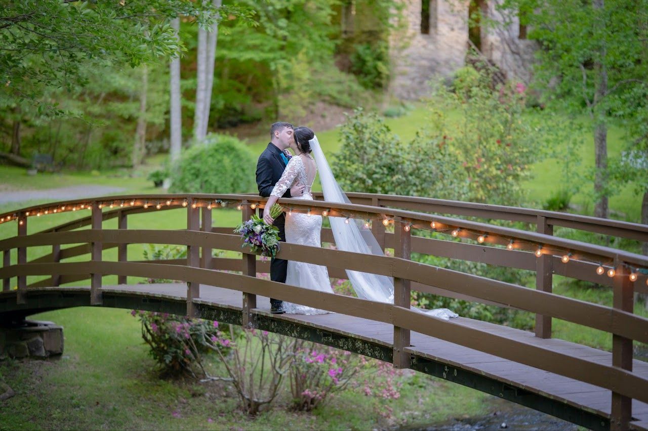 Bride and groom kissing on a wooden bridge, surrounded by greenery, with a castle in the background.