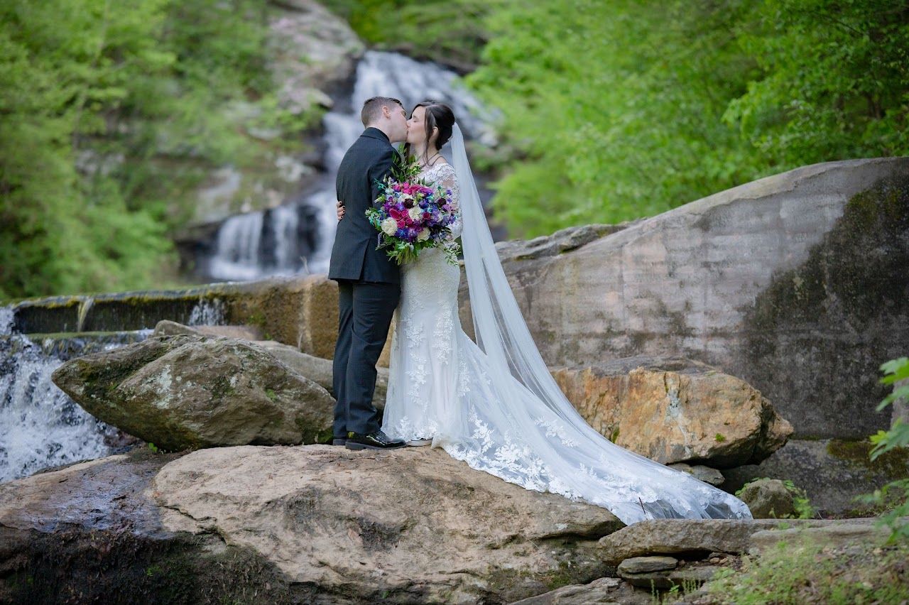 Bride and groom kissing near a waterfall, she wears a white lace gown, he wears a dark suit.