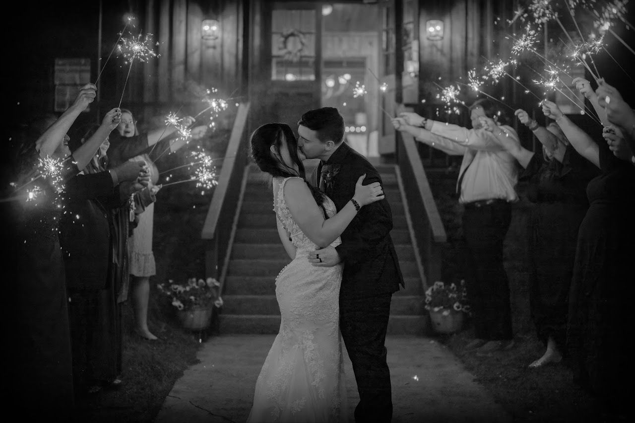 Newlyweds kiss amidst sparklers, flanked by wedding party, at a venue entrance at night.