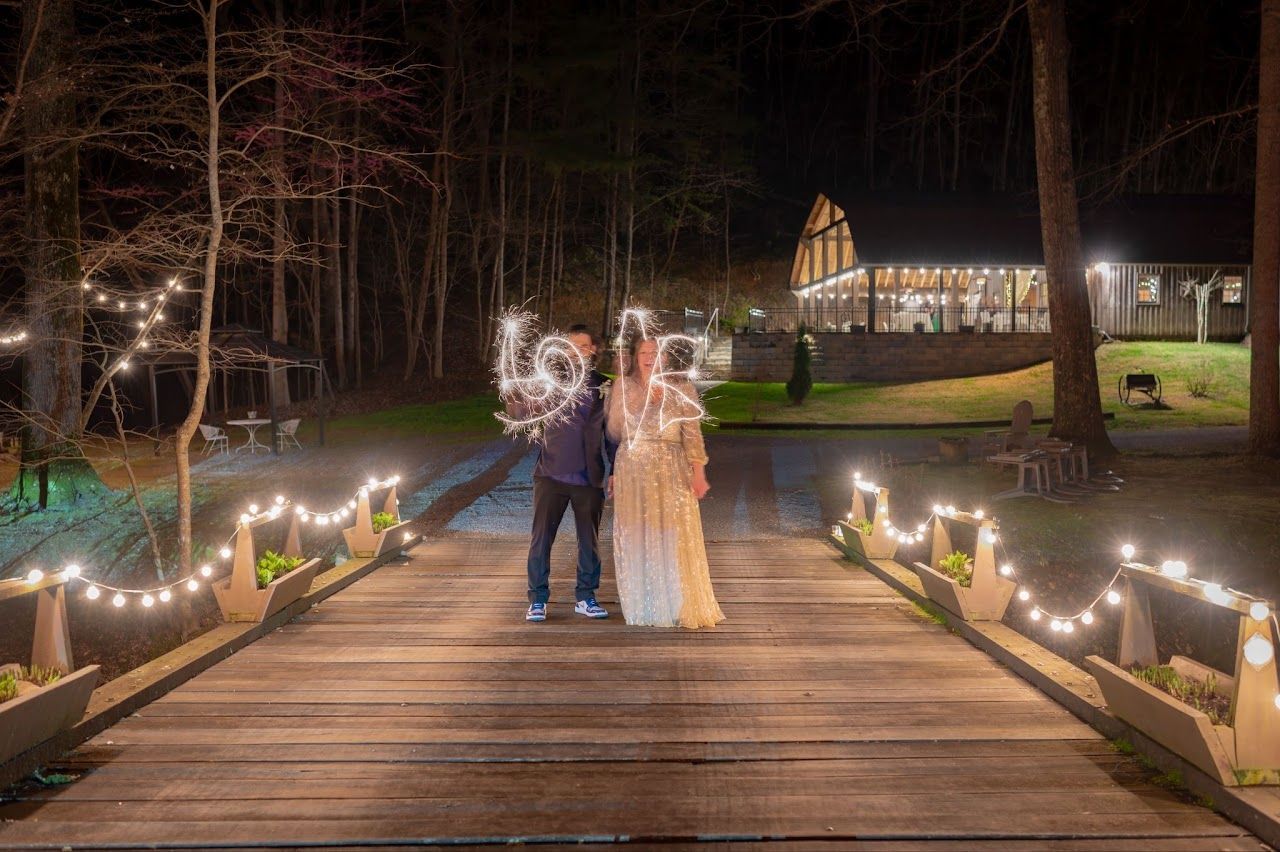 Couple on a wooden bridge, holding sparklers. Lit by string lights in front of a cabin at night.