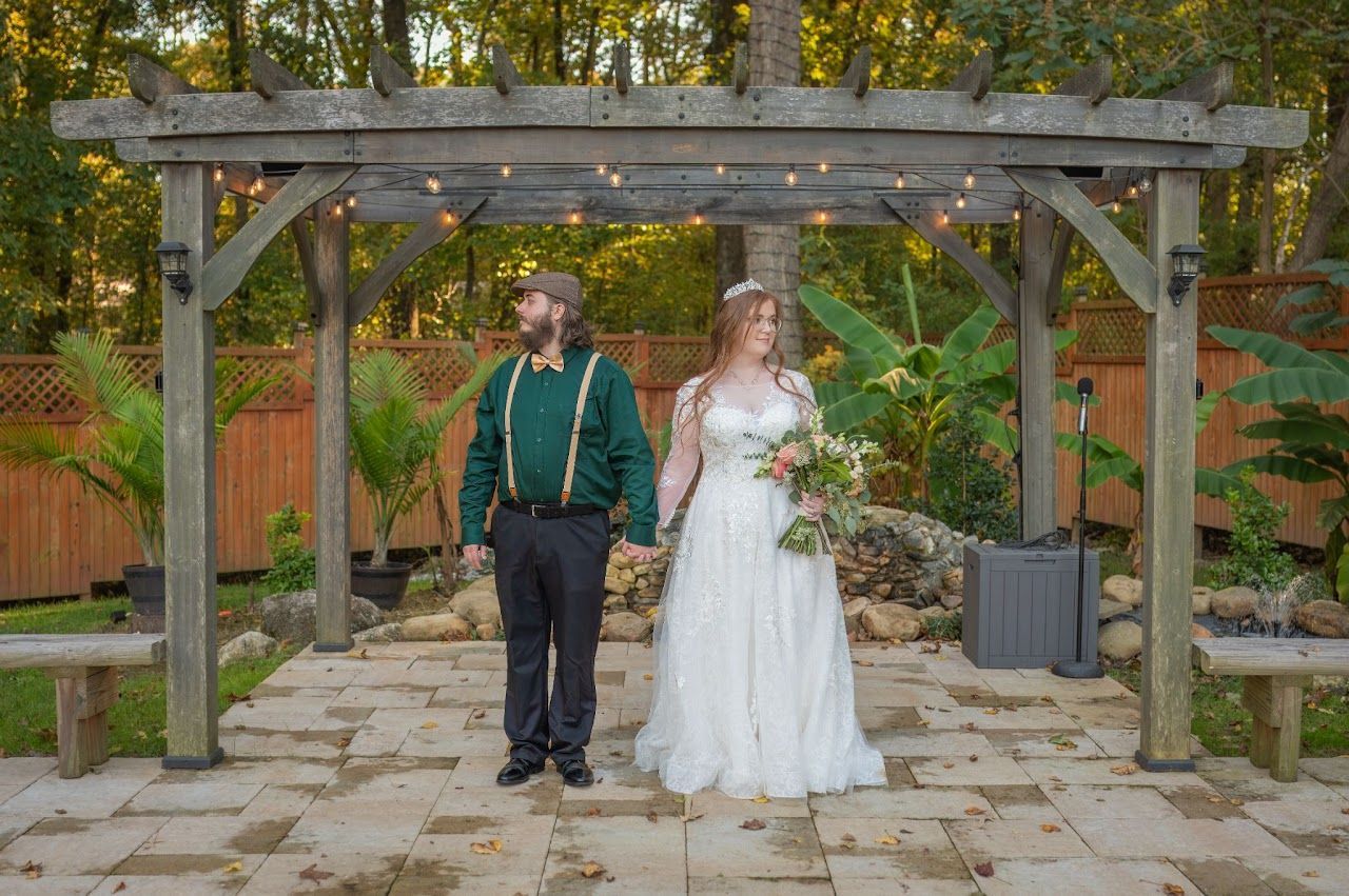 Bride and groom holding hands under a wooden arbor. Bride wears a white gown. Groom wears green shirt and suspenders.