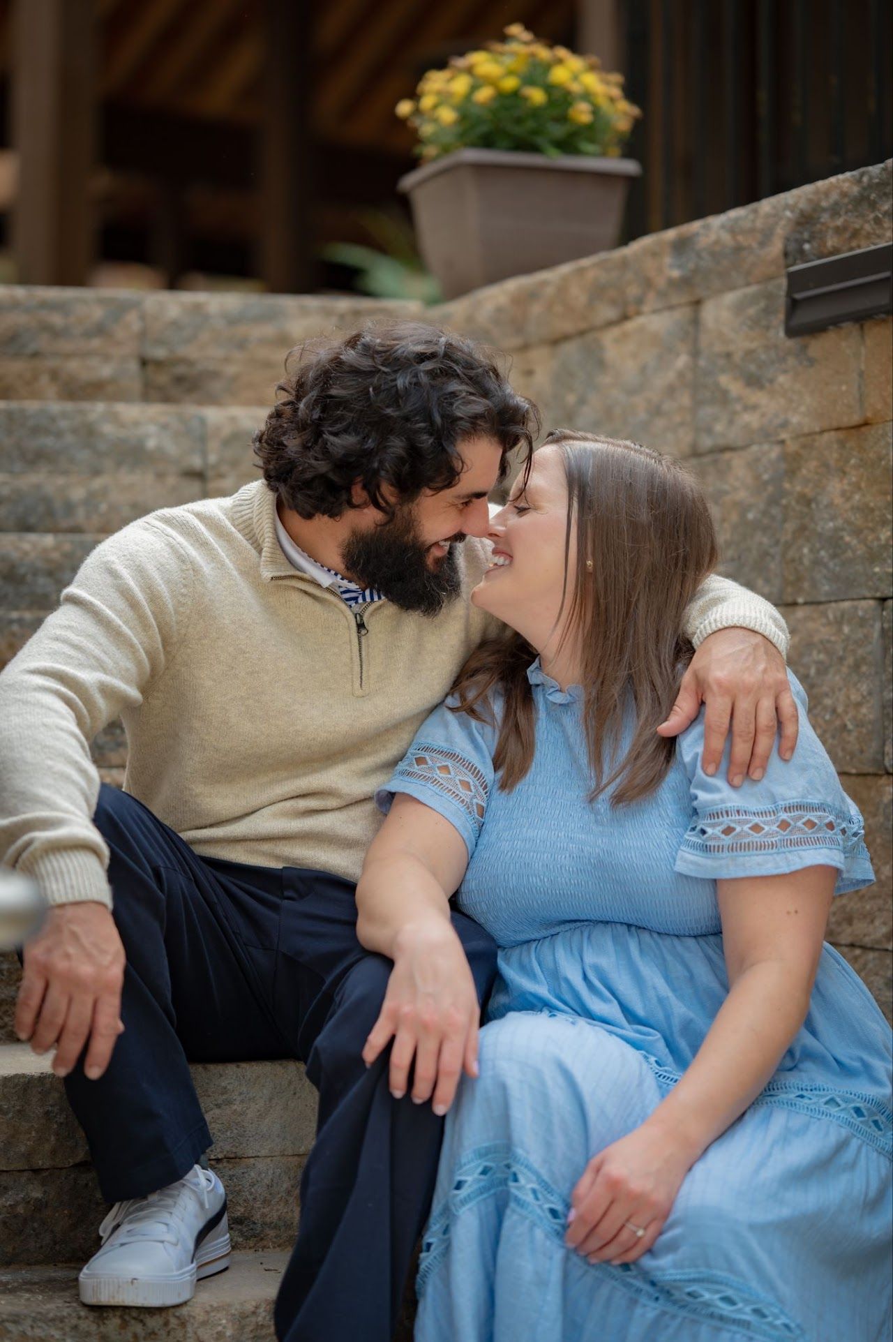Couple about to kiss on stone steps; man with beard, woman in blue dress, flowers in background.