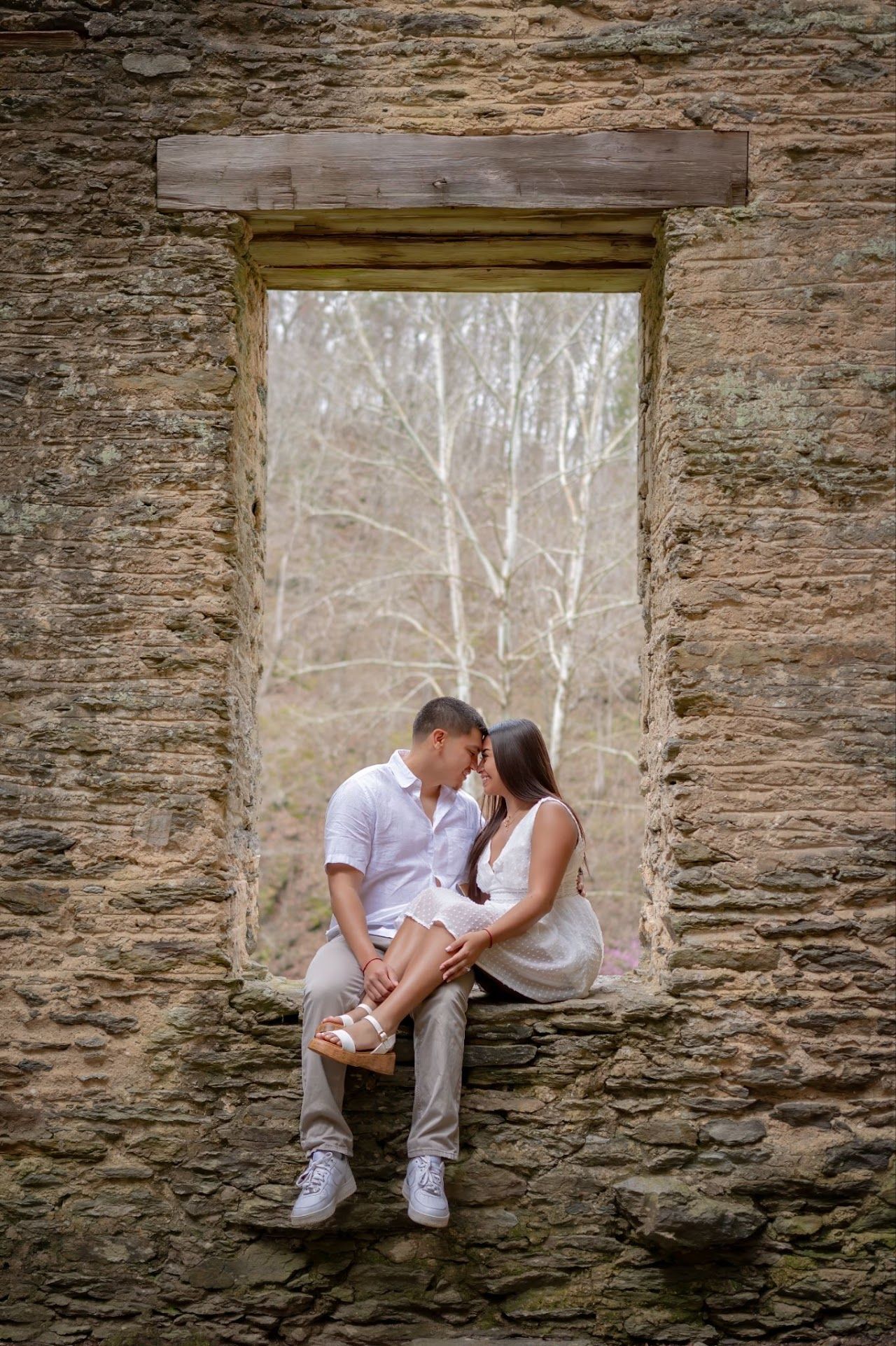 Couple embracing in a window frame of a stone building. Trees in the background.
