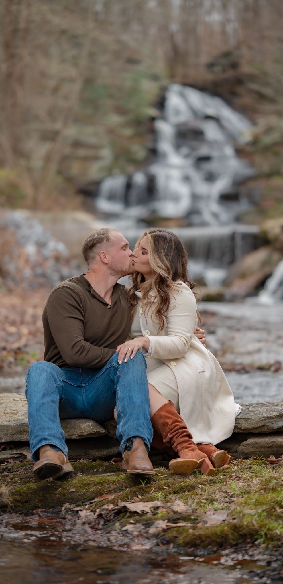 Couple kissing by a waterfall. Man in jeans and brown shirt. Woman in white dress and brown boots. Outdoors.