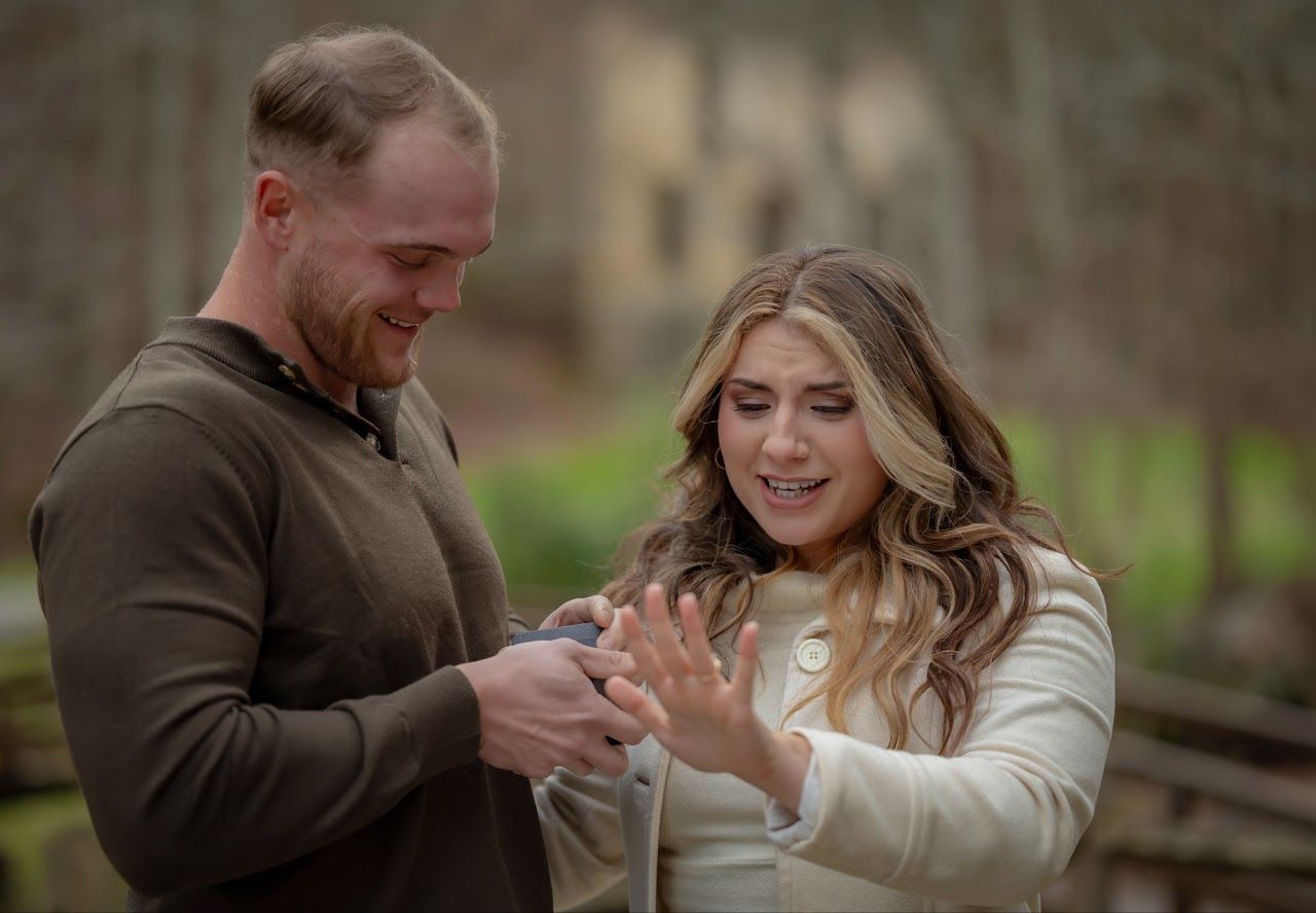 Man proposing to woman outdoors, holding ring box. Woman looks surprised and excited.