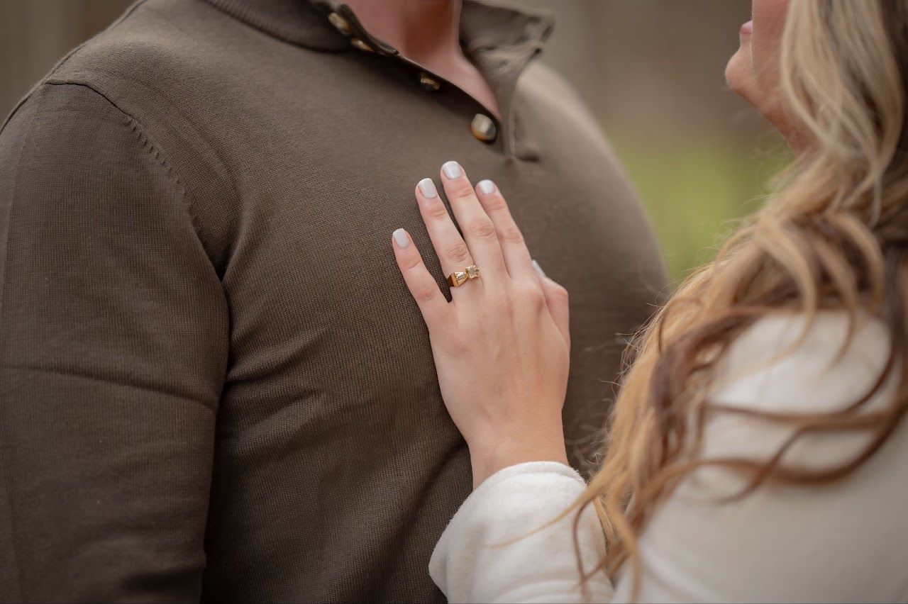 Woman's hand with ring on man's chest. Man wears brown shirt; woman's blonde hair visible. Outdoors.
