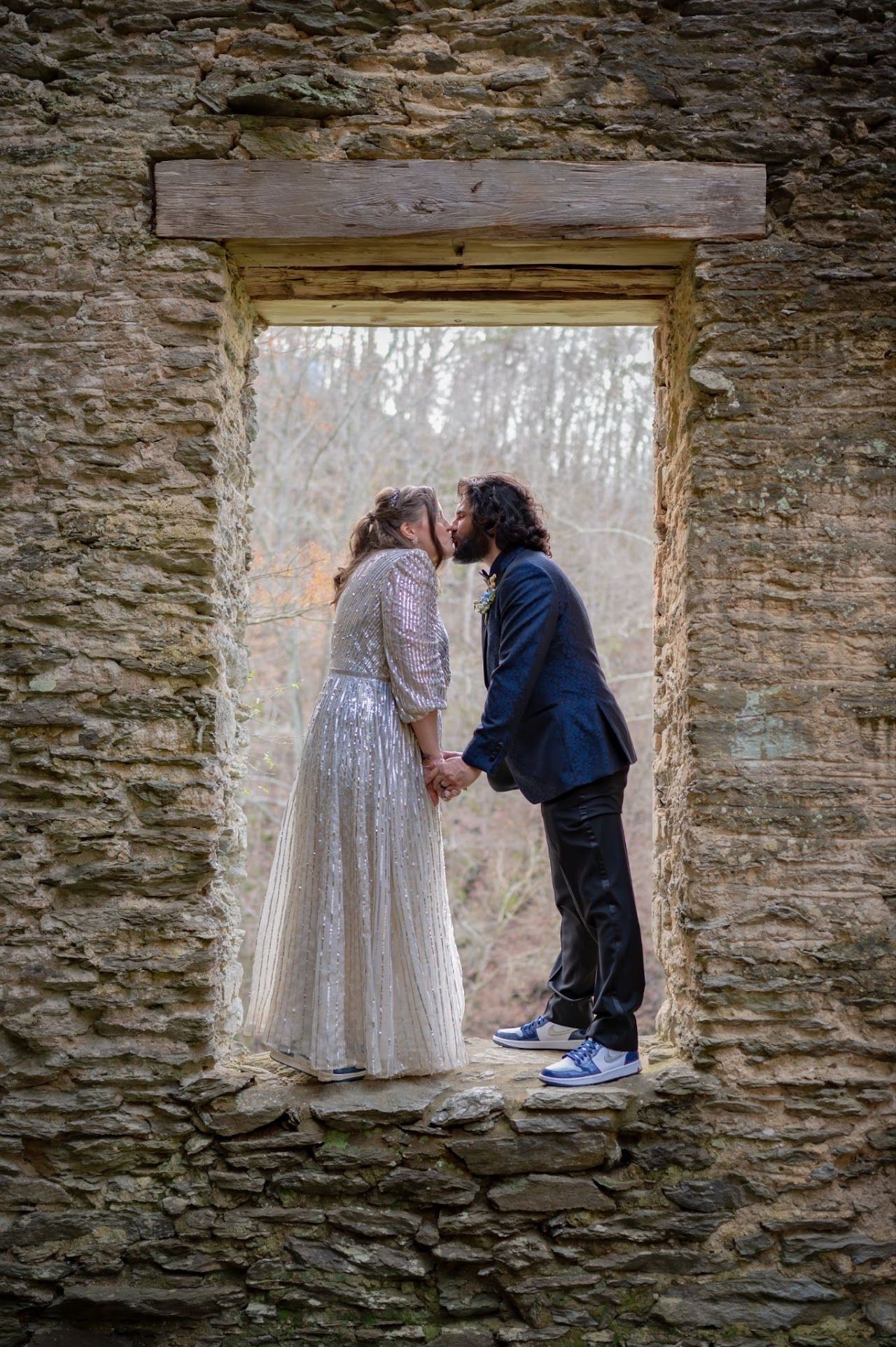 Couple kissing in an old stone window, holding hands. Woman in sparkly gown, man in navy jacket and sneakers.