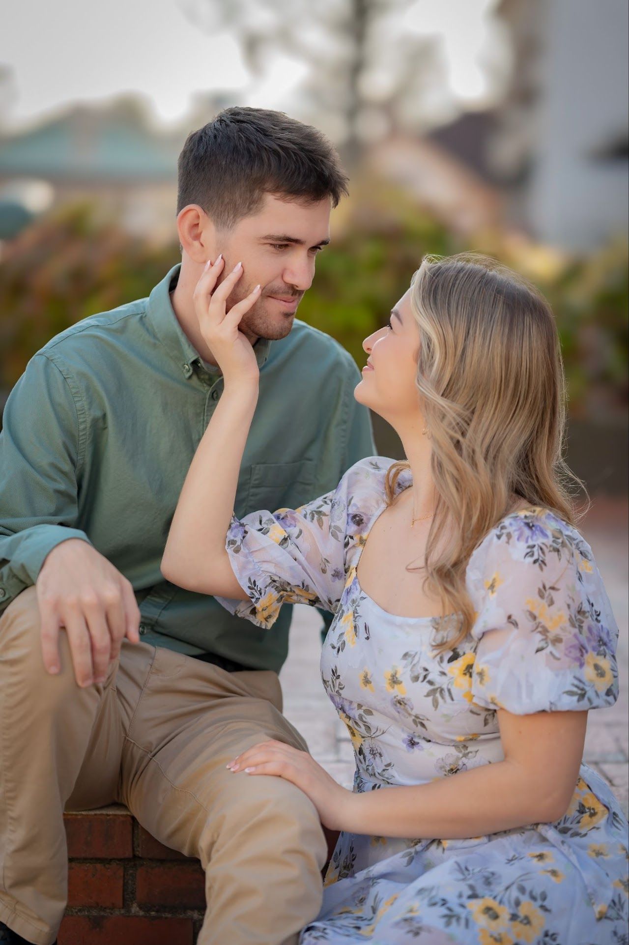 Couple gazing lovingly at each other; woman touching man's face. Outdoors; woman in floral dress, man in green shirt.