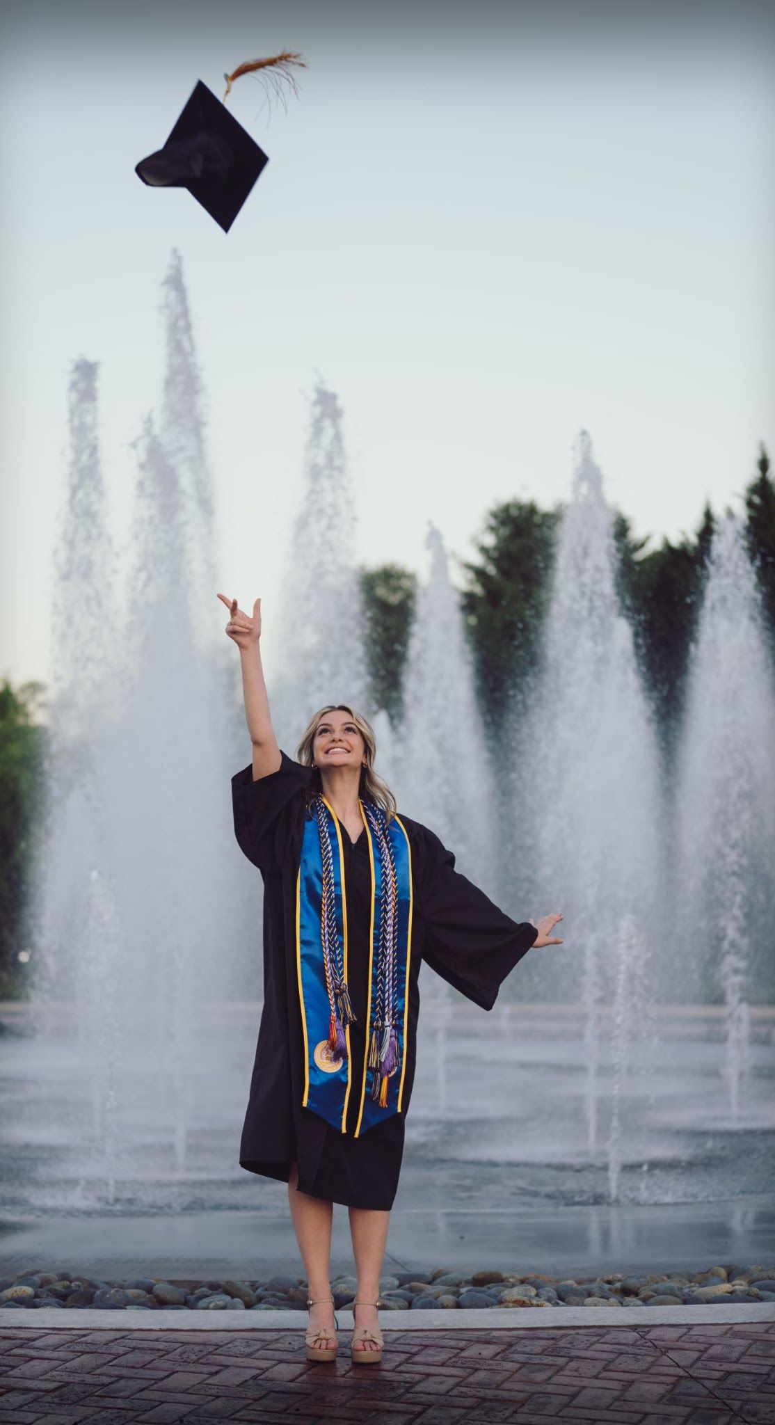 Woman in graduation gown tossing cap in the air near a fountain.