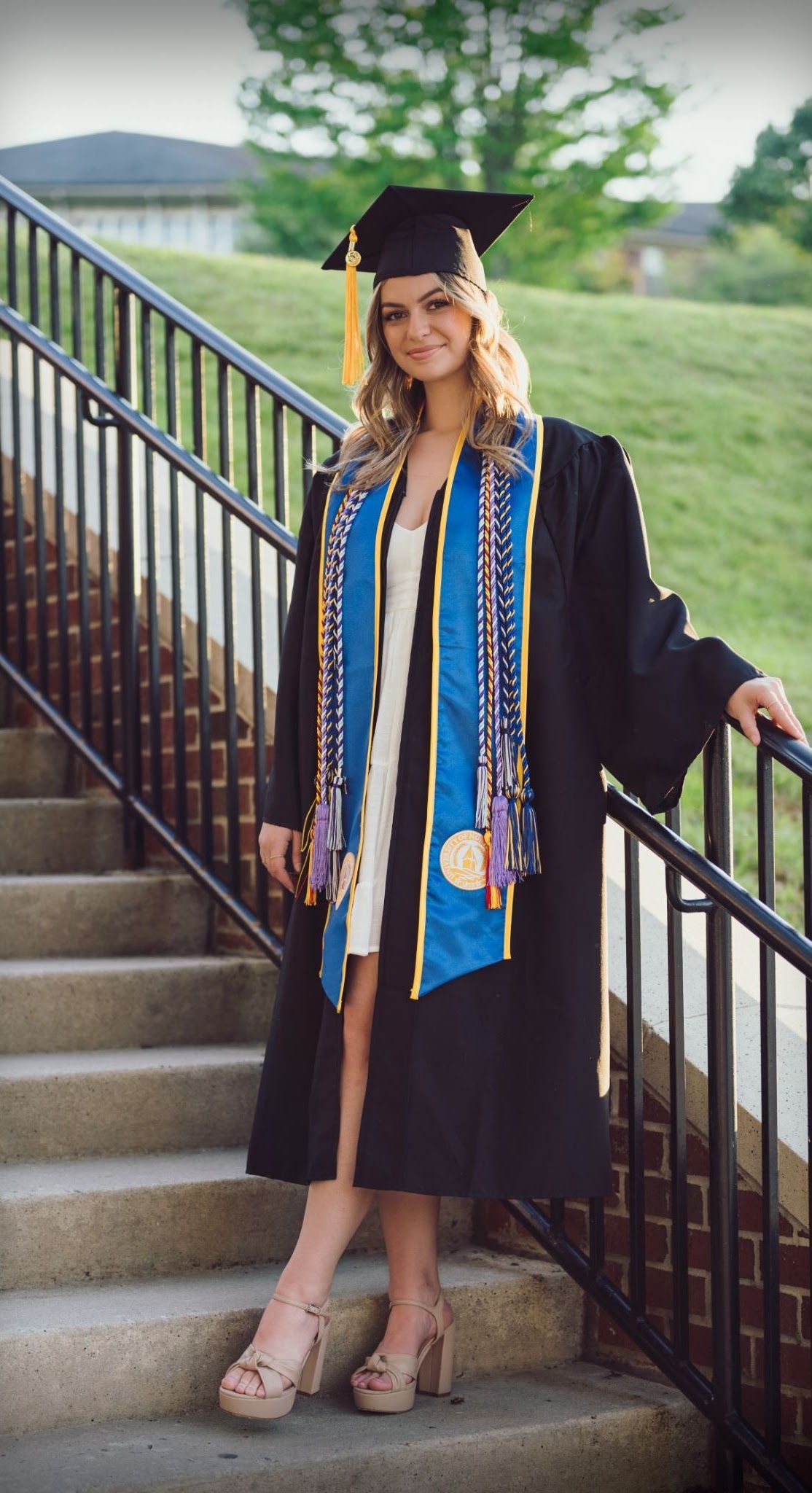 Woman in graduation gown and cap stands on stairs, smiles. Blue, white, black, gold colors.