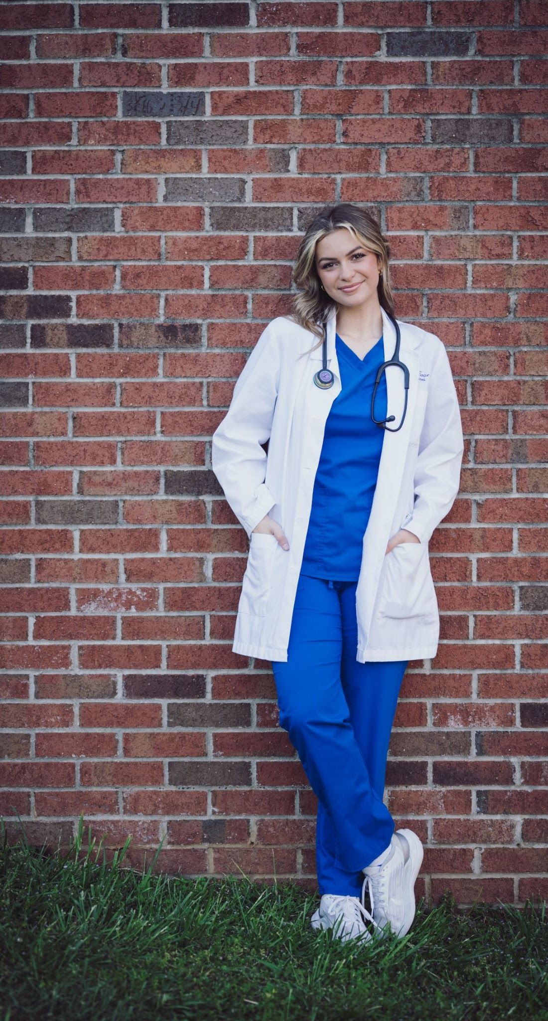 Woman in blue scrubs and white coat leaning against brick wall, stethoscope around neck.