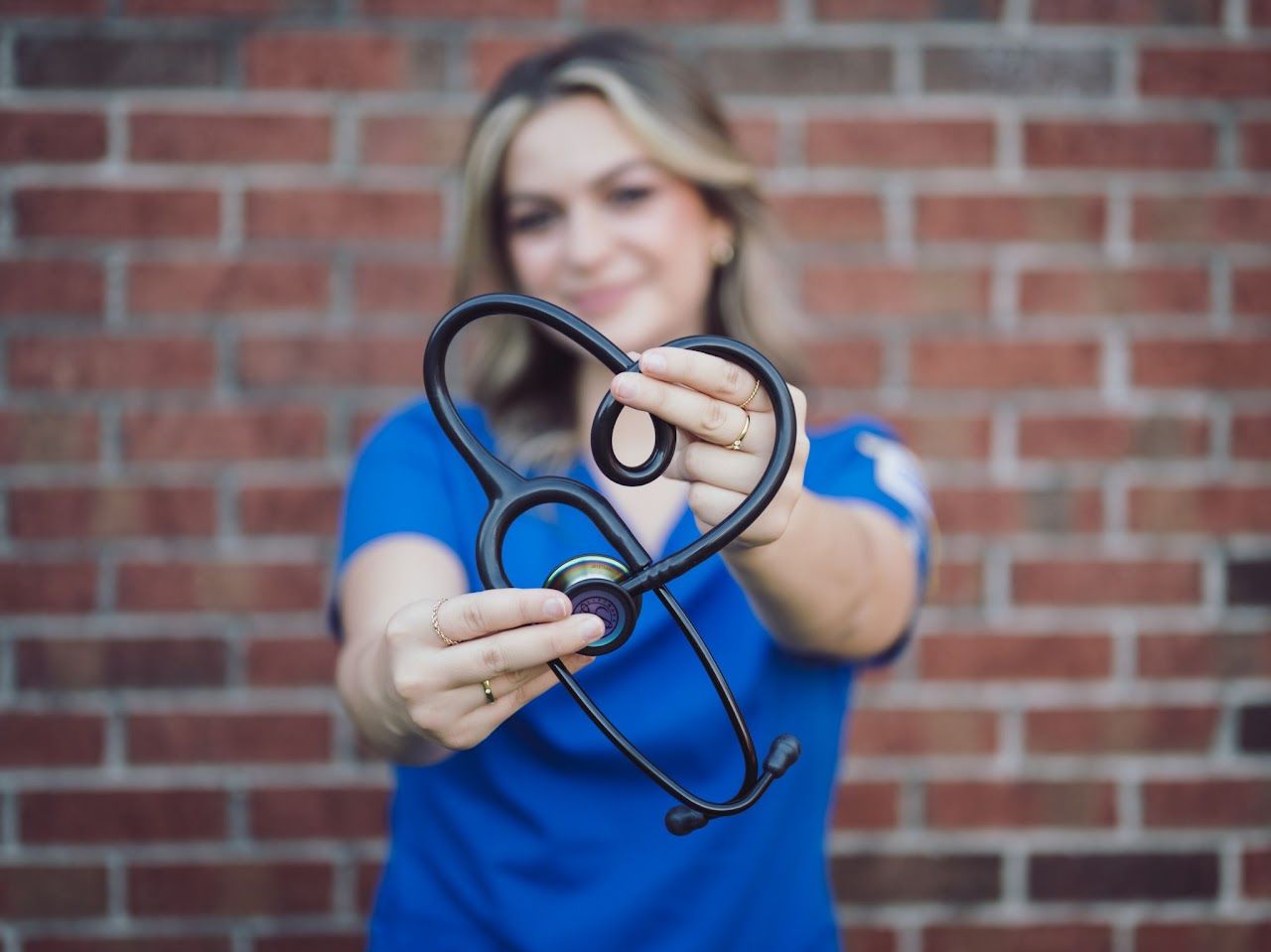 Nurse in blue scrubs holding stethoscope in heart shape against a brick wall.