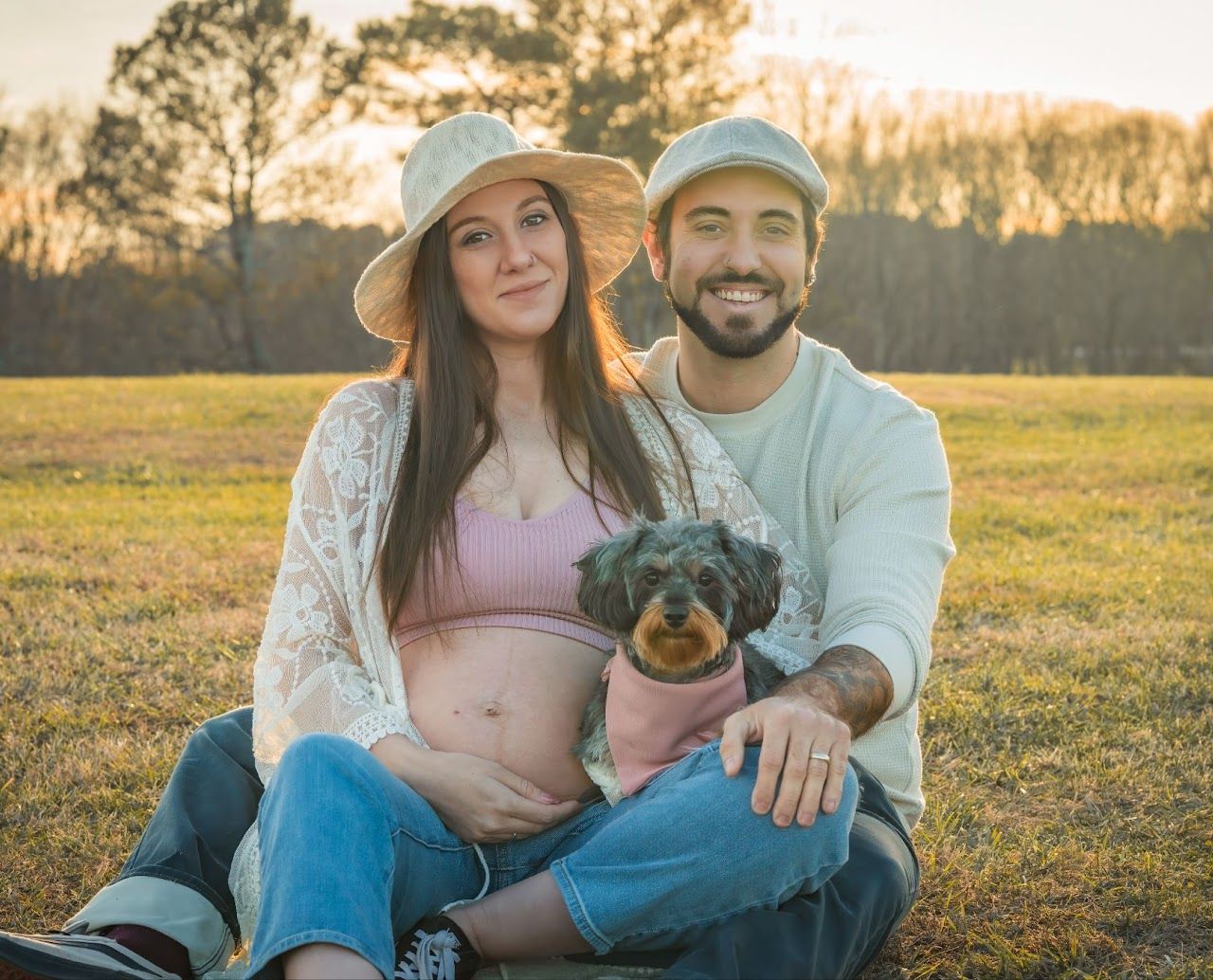 Pregnant couple and dog sitting on grass. Woman holds belly. Man smiles; sunset.