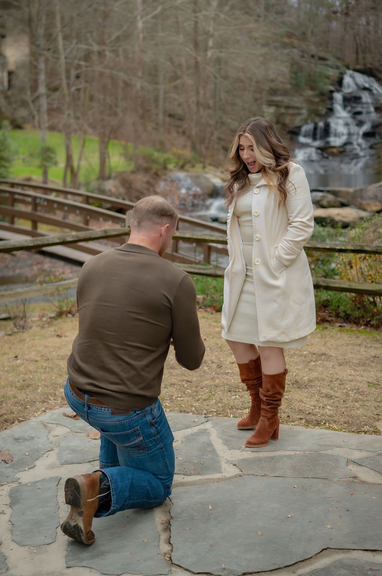 Man on one knee proposing to woman near a waterfall.