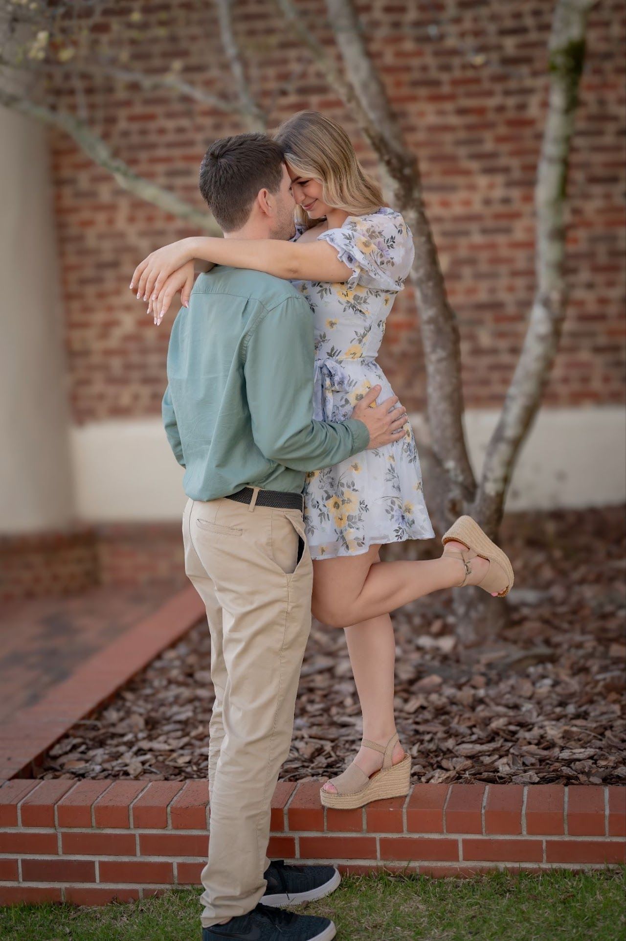 Man holding woman in a hug near brick wall. She's wearing a floral dress and he's in a green shirt and khakis.