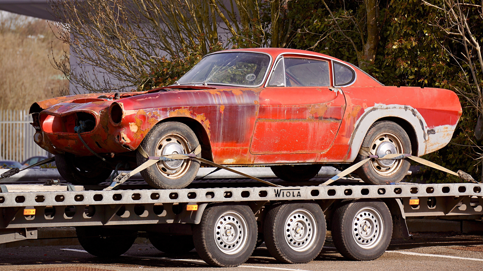 Red, rusty classic car on a flatbed trailer, wheels secured with straps.