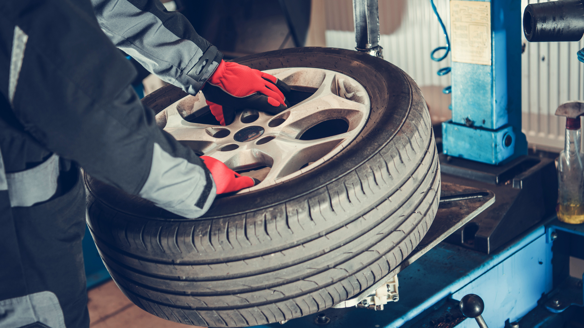 Mechanic in red gloves working on a car tire, near a blue machine, inside a garage.