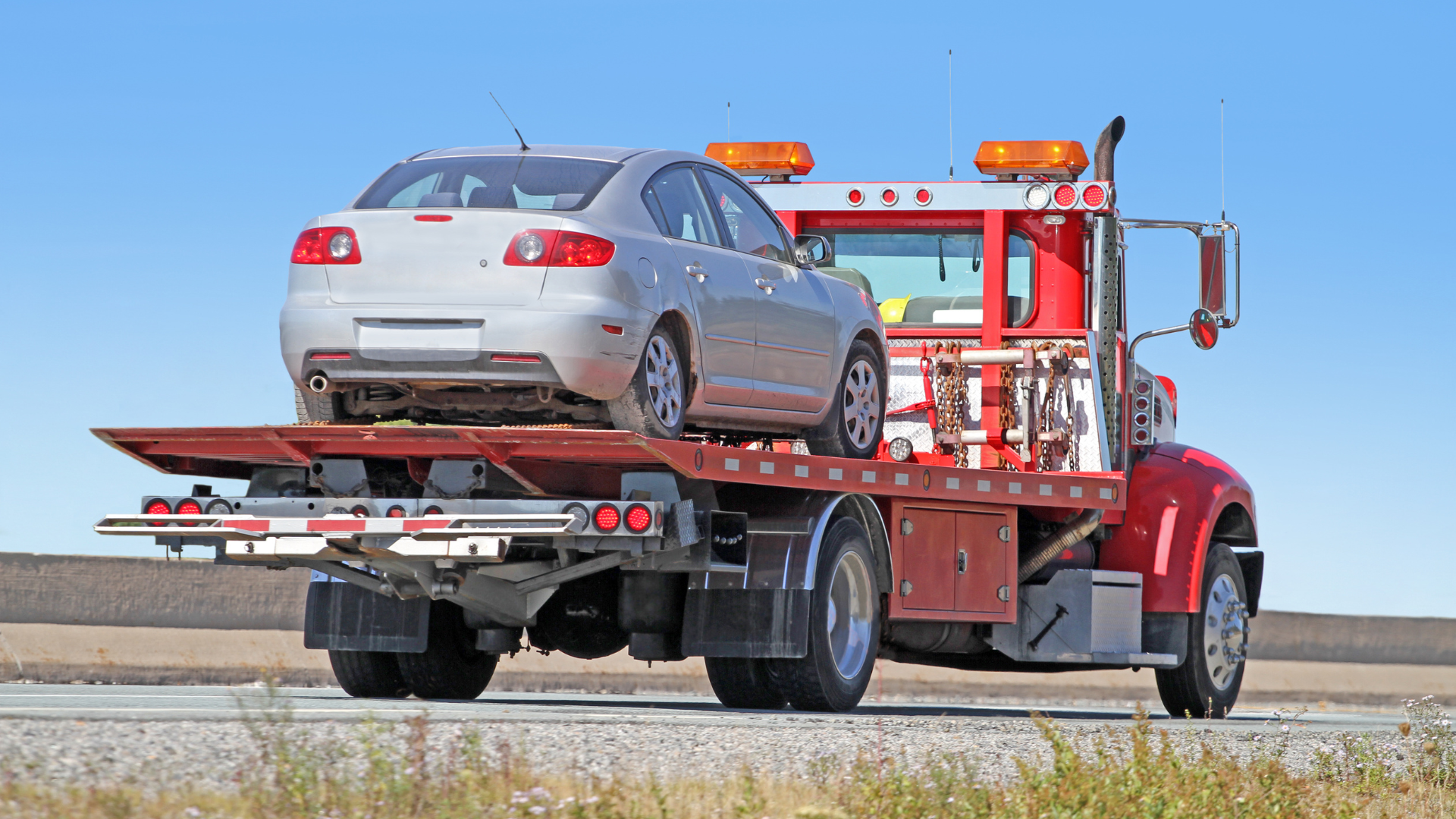 Silver car being towed on a red flatbed tow truck on a highway under a blue sky.