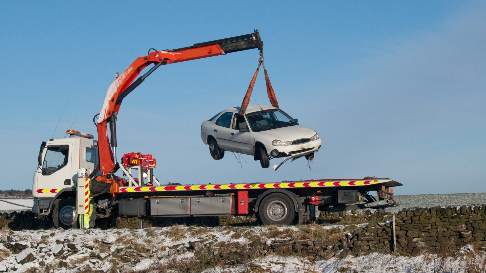 Tow truck lifting a damaged white car in a snowy field on a sunny day.