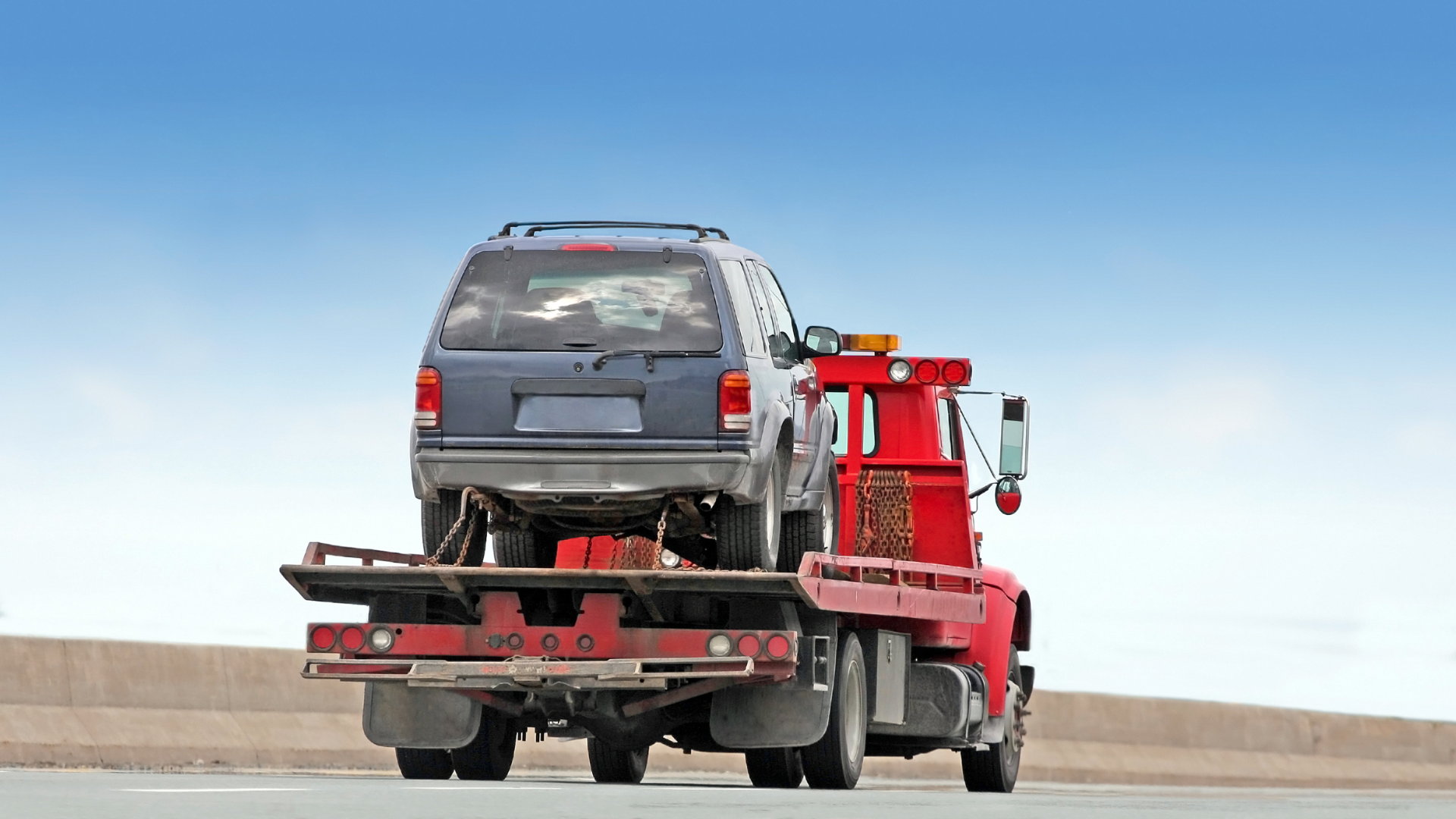 Blue SUV being towed on a red tow truck on a highway with blue sky.