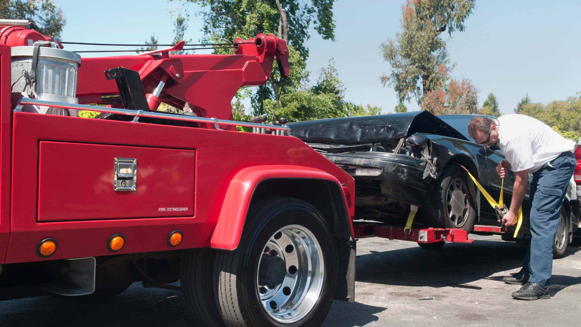 Red tow truck loading a damaged black car; a man secures straps outdoors.