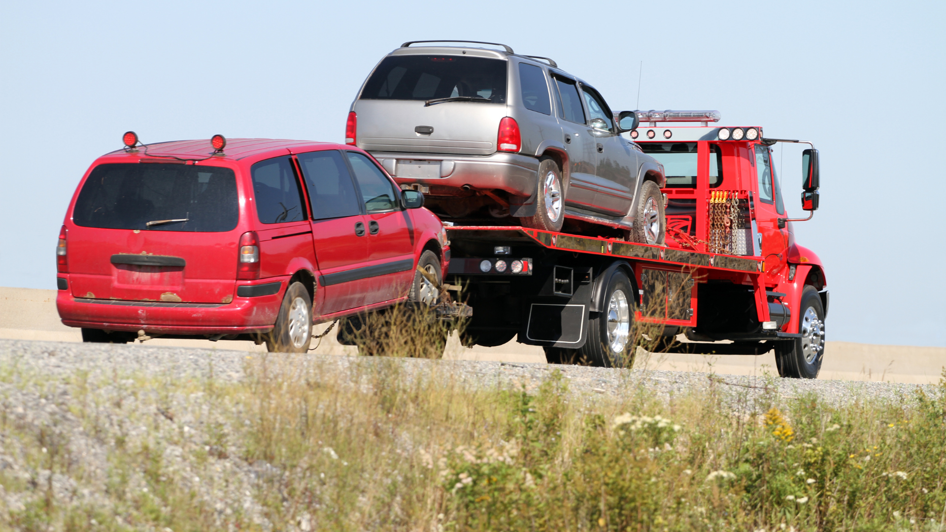Red tow truck carrying a silver SUV and a red minivan on a highway.