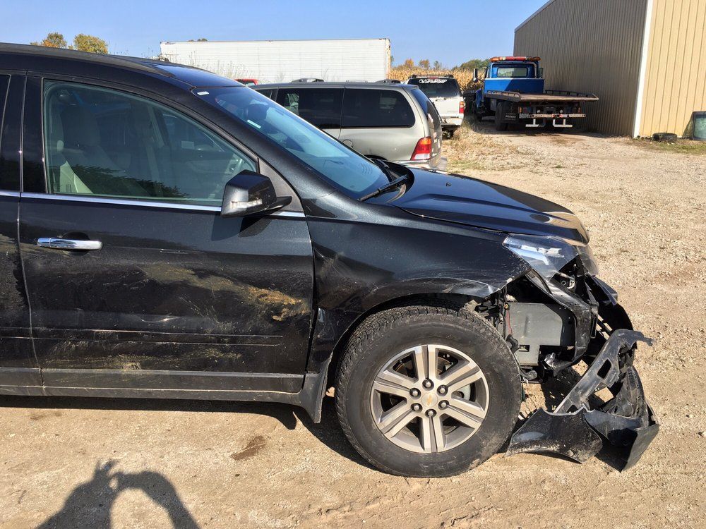 Dark SUV with front-end damage, missing bumper and headlight, parked outdoors on a sunny day.