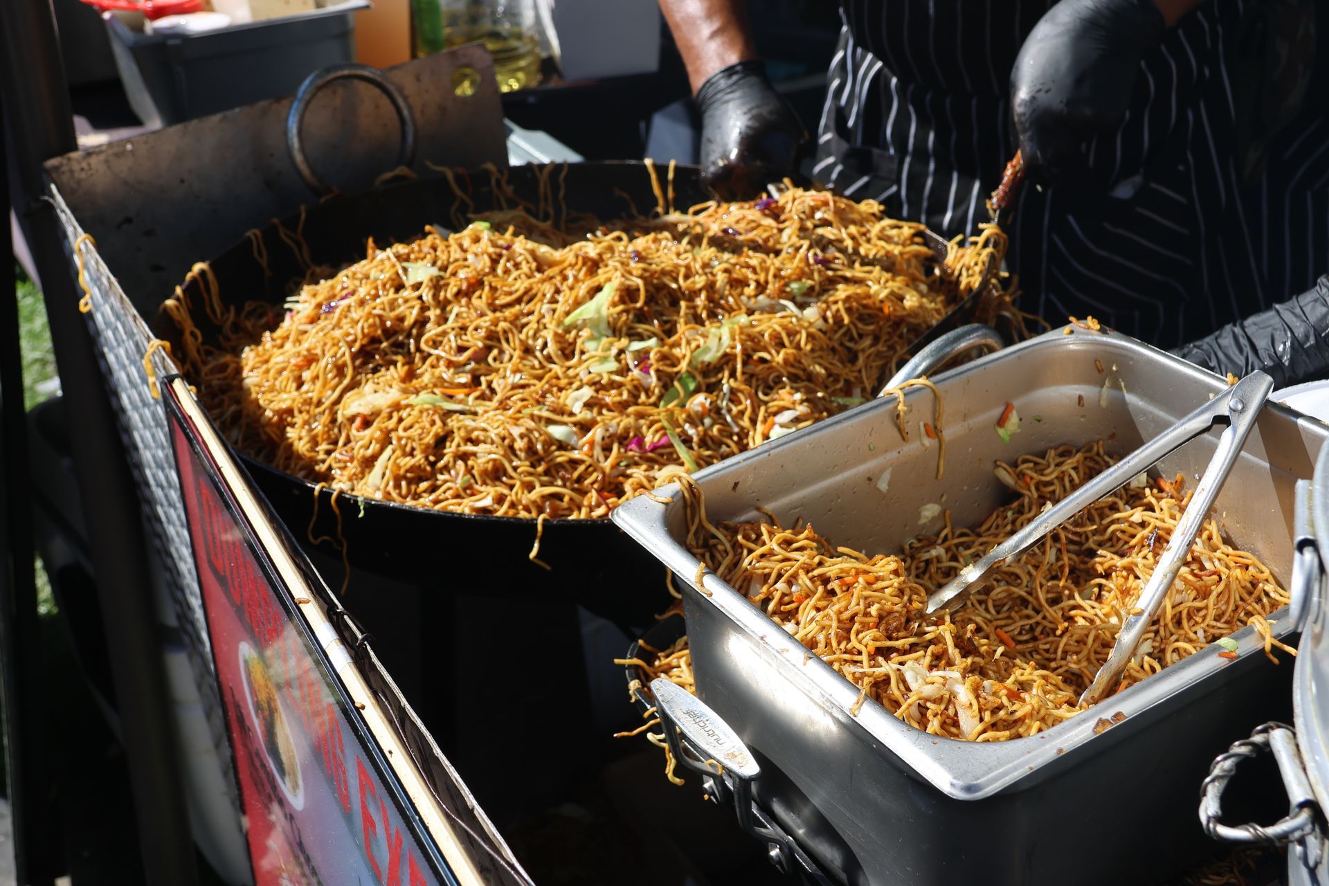 A large wok full of noodles at a food stall. A person stirs the noodles.