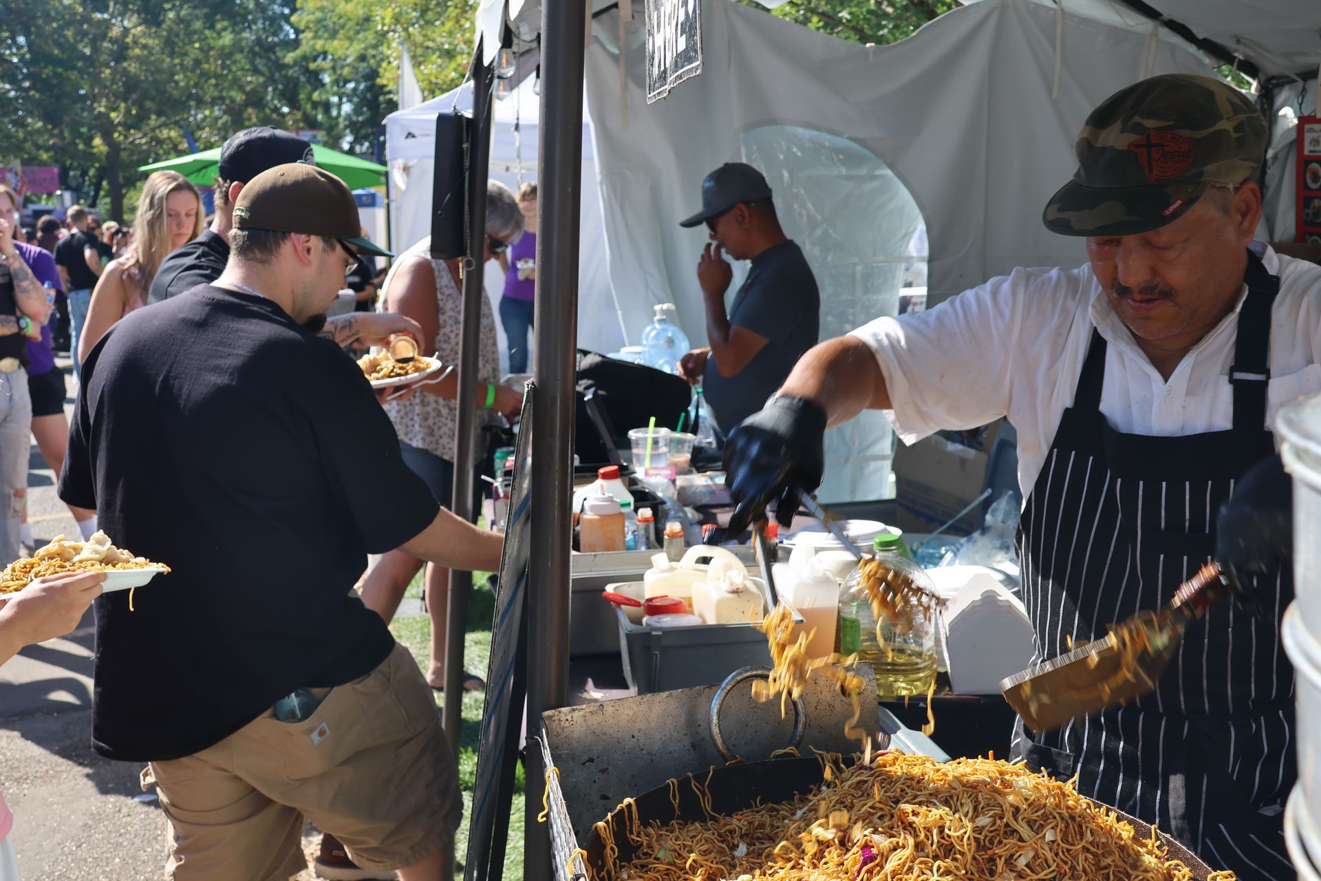 Chef cooking food at a food stall outdoors. Customers wait in line.