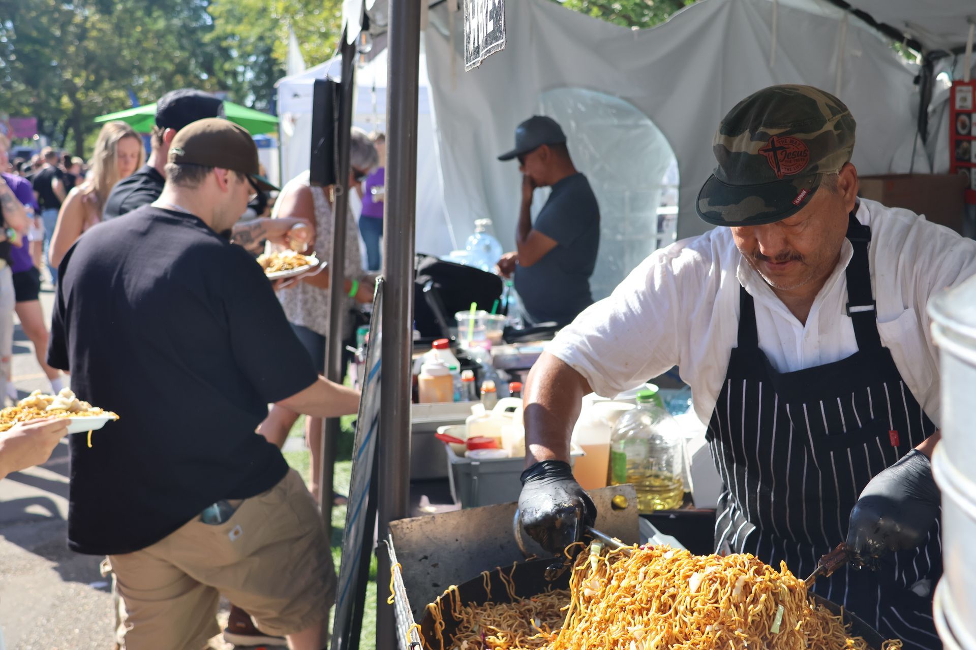 Man cooking noodles at a food stall; customers waiting. Outdoor setting, sunny, and busy.