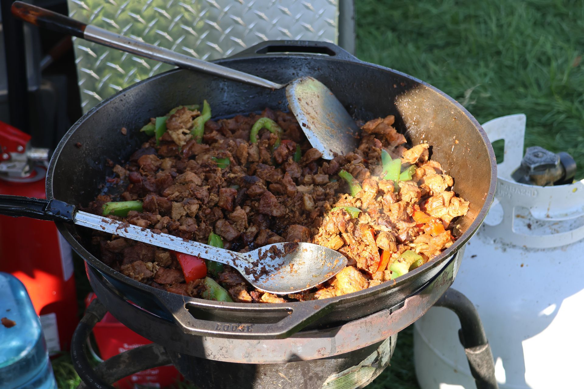 Sausage and peppers cooking in a large cast iron skillet over a propane burner outdoors.