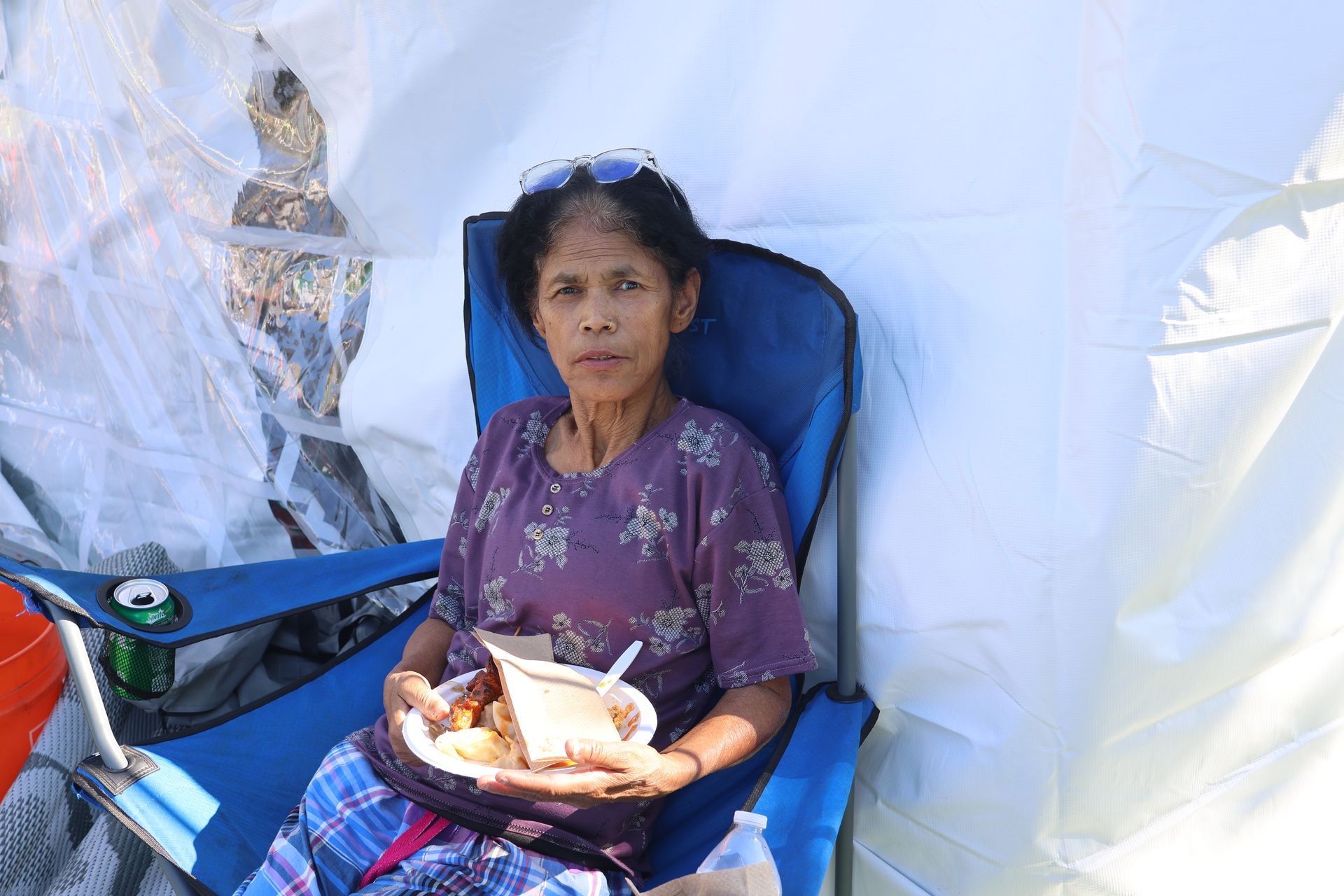 Woman in purple shirt, seated, eating. Outdoors, near a white tent.