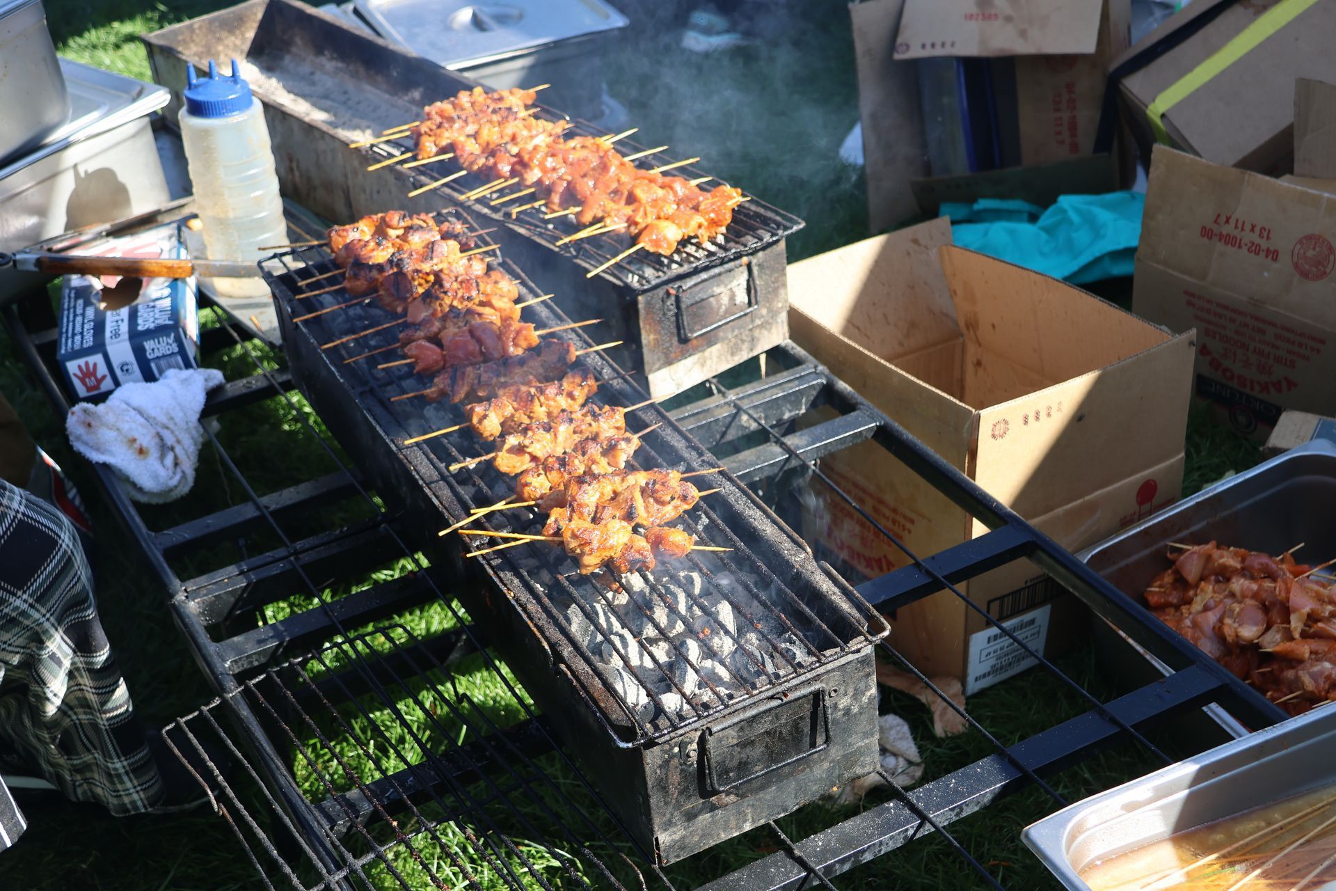 Barbecue skewers grilling over charcoal on outdoor grates; boxes nearby.