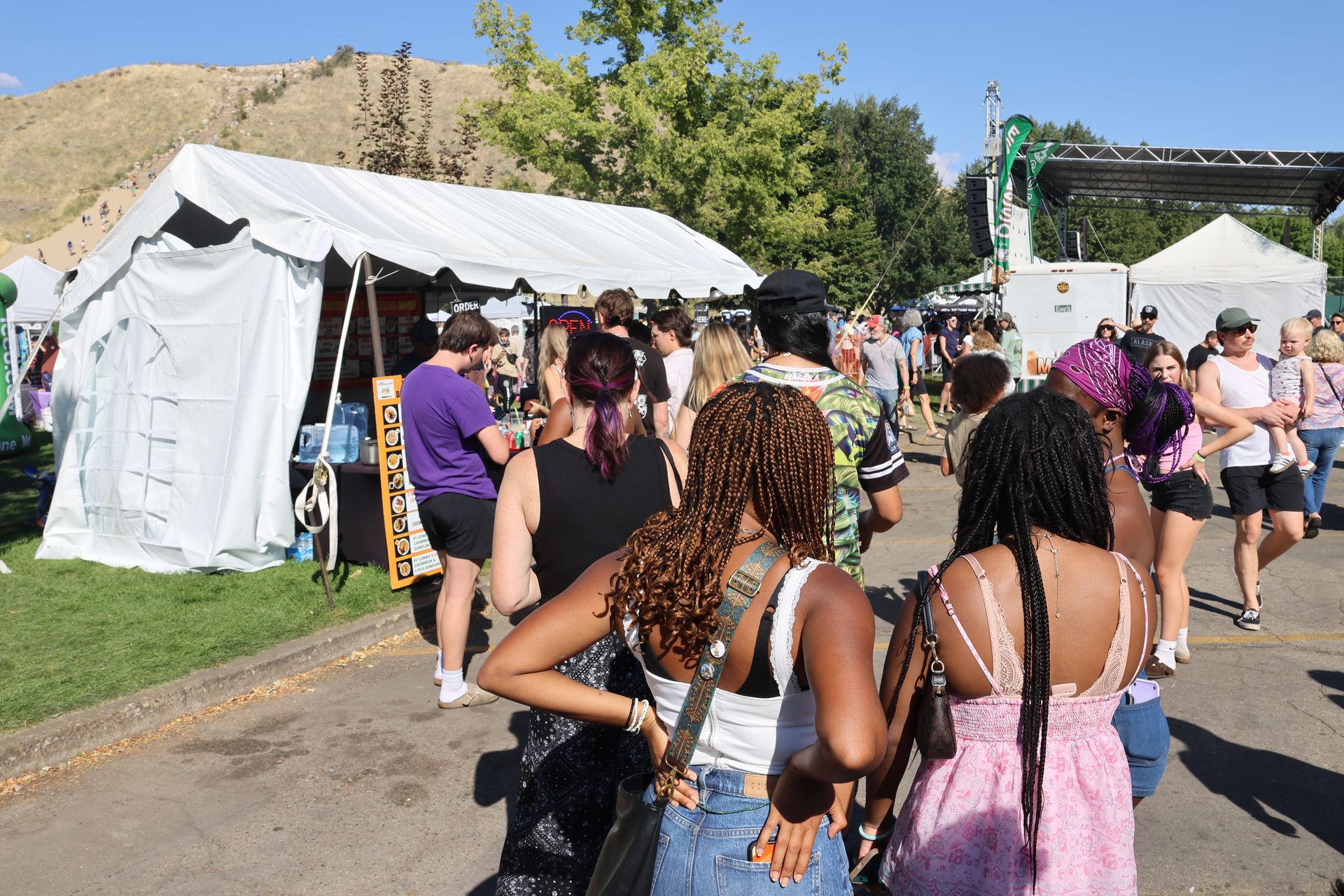 People at outdoor festival, walking past tents and stages on a sunny day.
