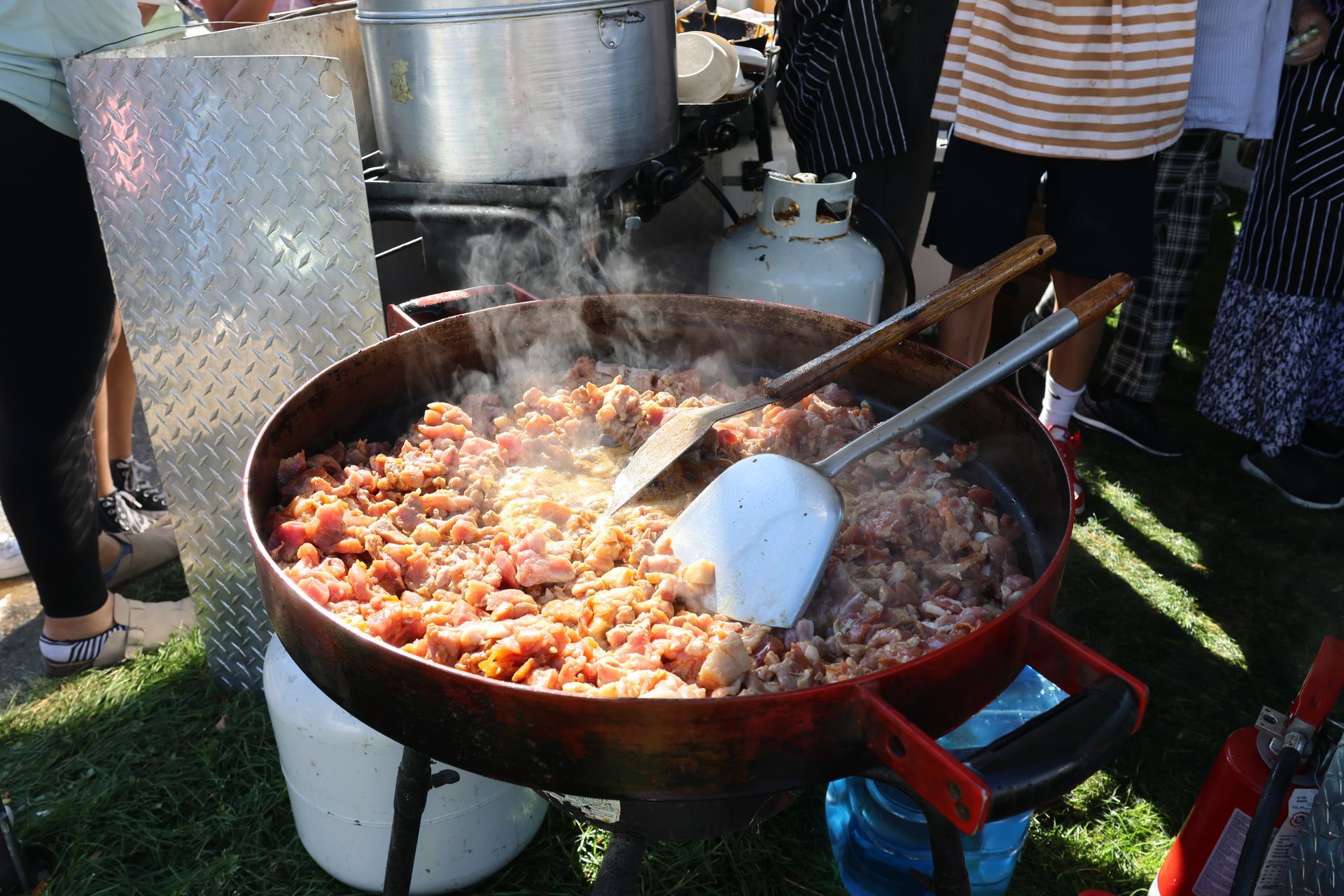 Large pan of cooking meat on a propane burner outdoors, with people nearby.